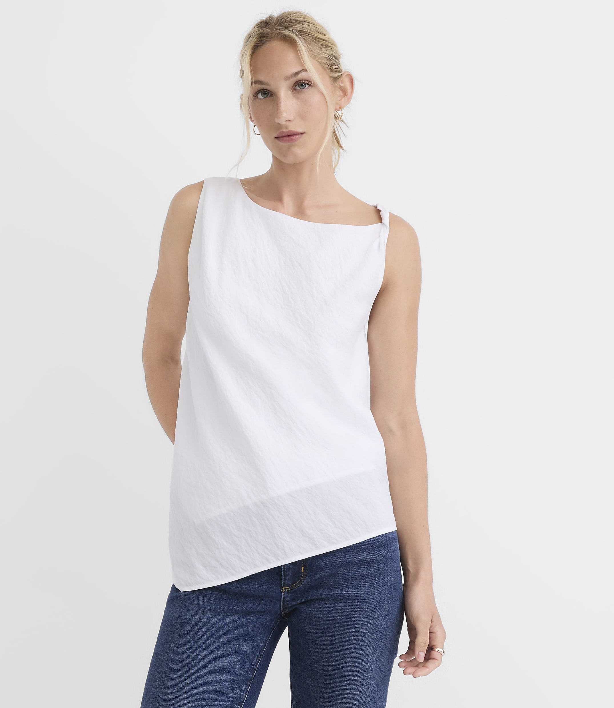 A woman in a white asymmetrical top and blue jeans stands against a plain white background.