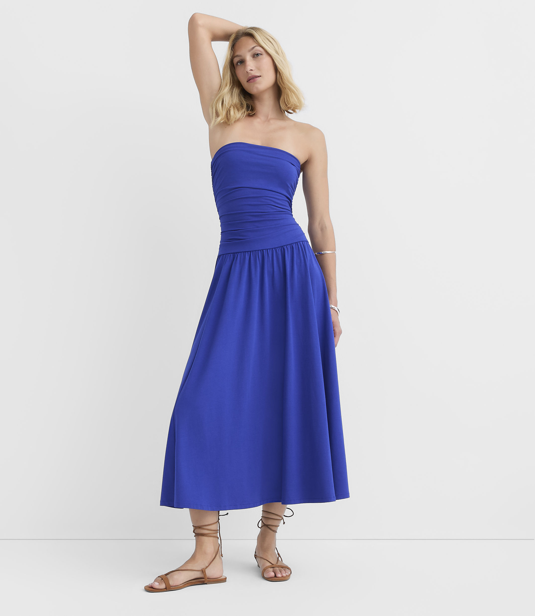 A woman in a blue strapless dress and sandals stands against a plain white background.