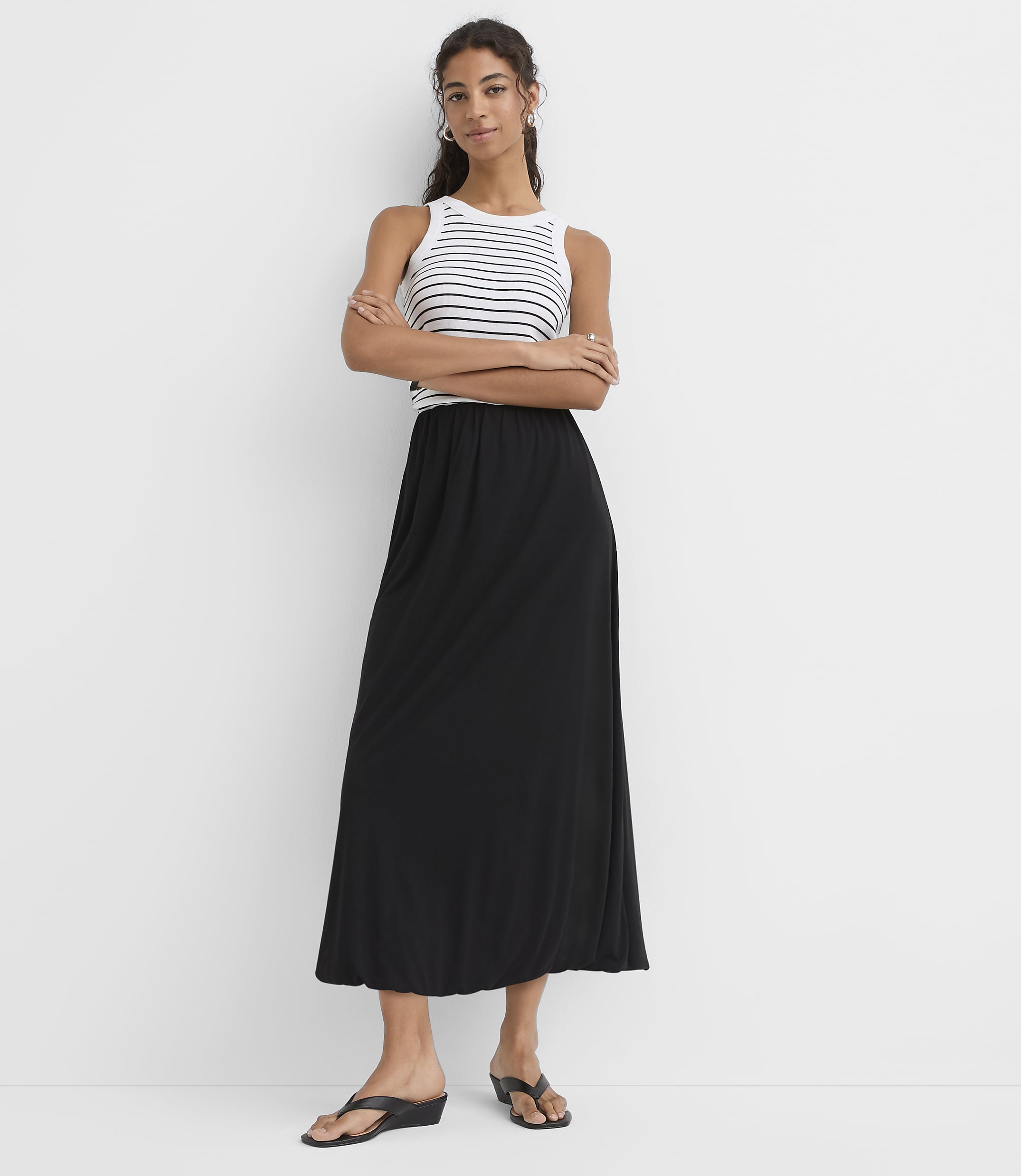 A woman in a striped tank top and black maxi skirt stands with her arms crossed against a plain white background.