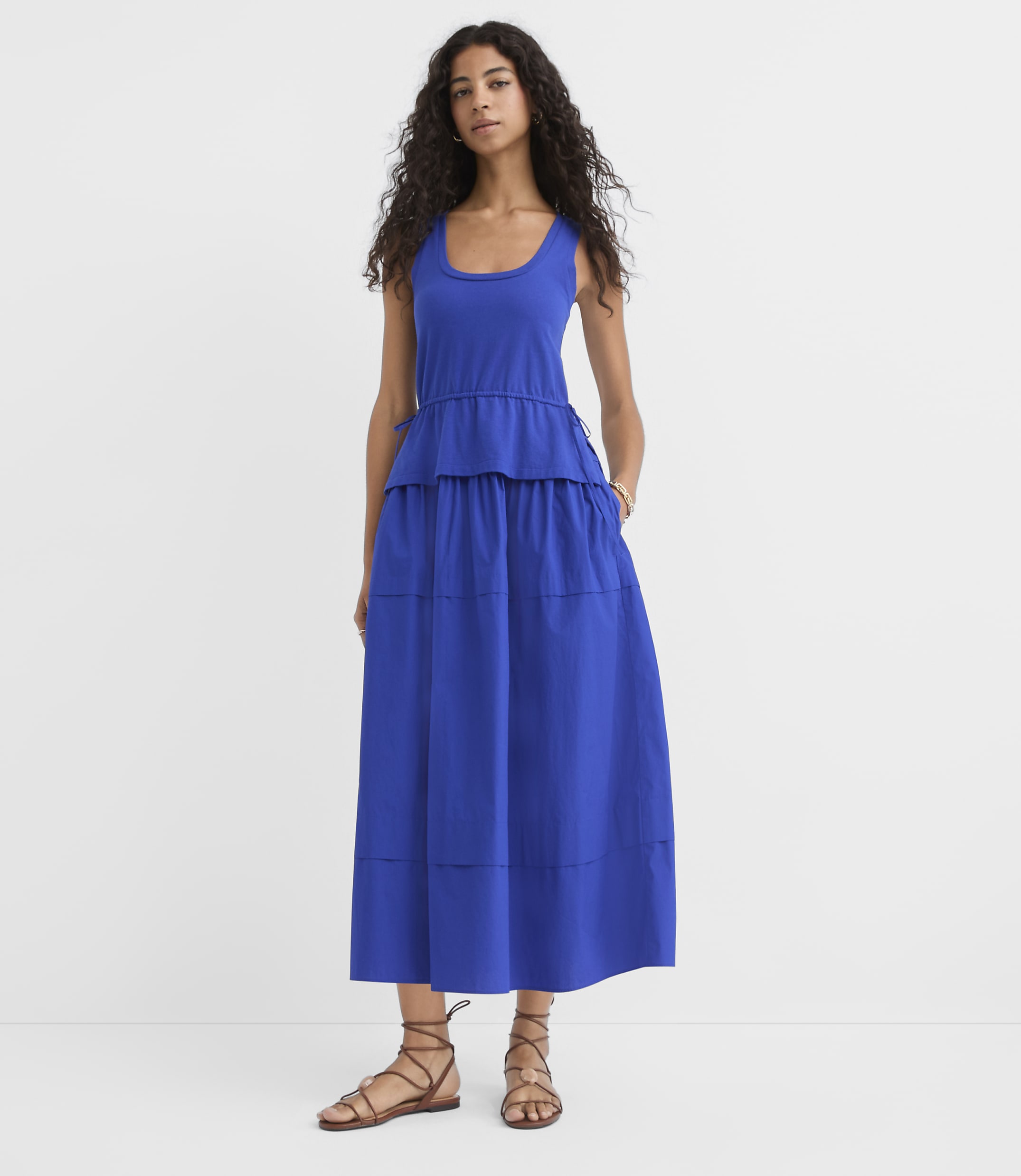 A woman in a blue tiered dress and sandals stands against a plain white background.