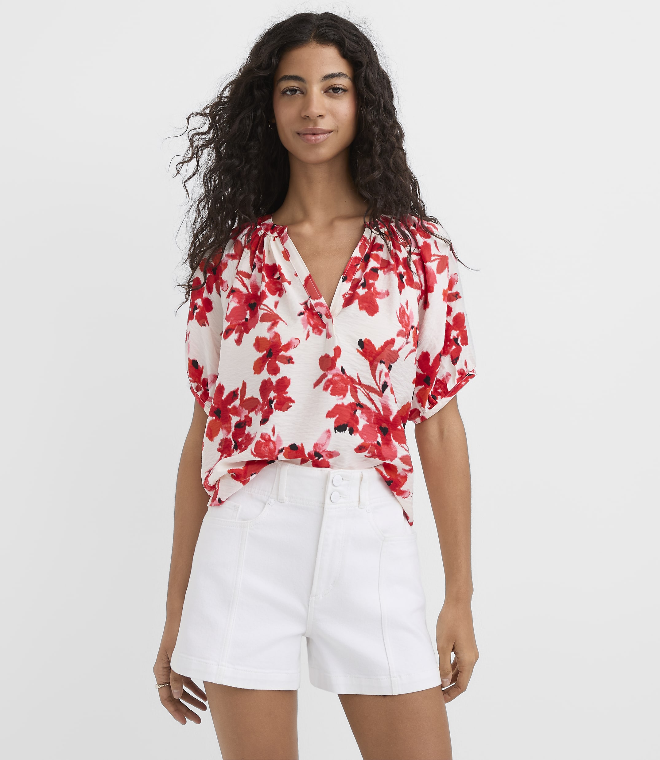 A woman in a floral blouse and white shorts stands against a plain white background.