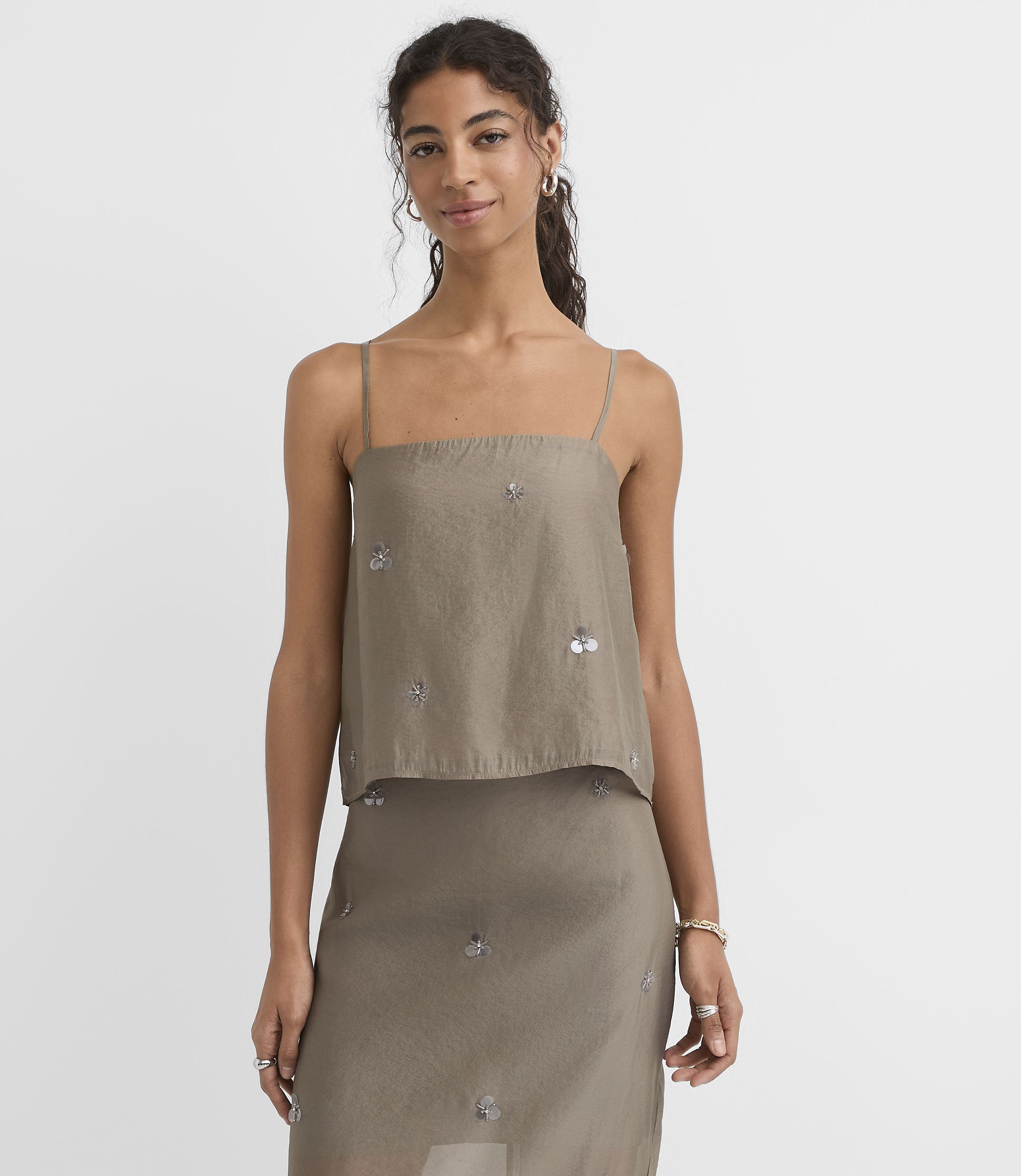 A woman in a taupe camisole top with sequin embellishments stands against a plain white background.