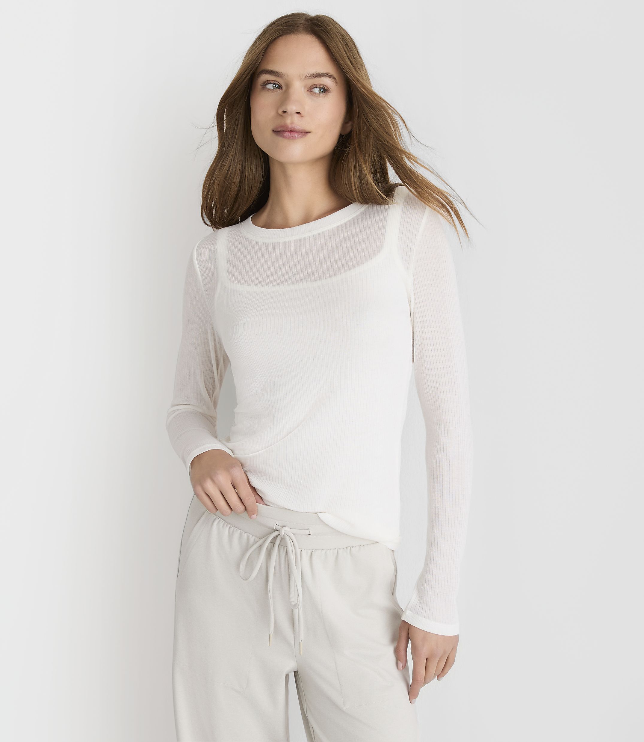 A woman in a white sheer long-sleeved top and light gray drawstring pants stands against a plain white background.