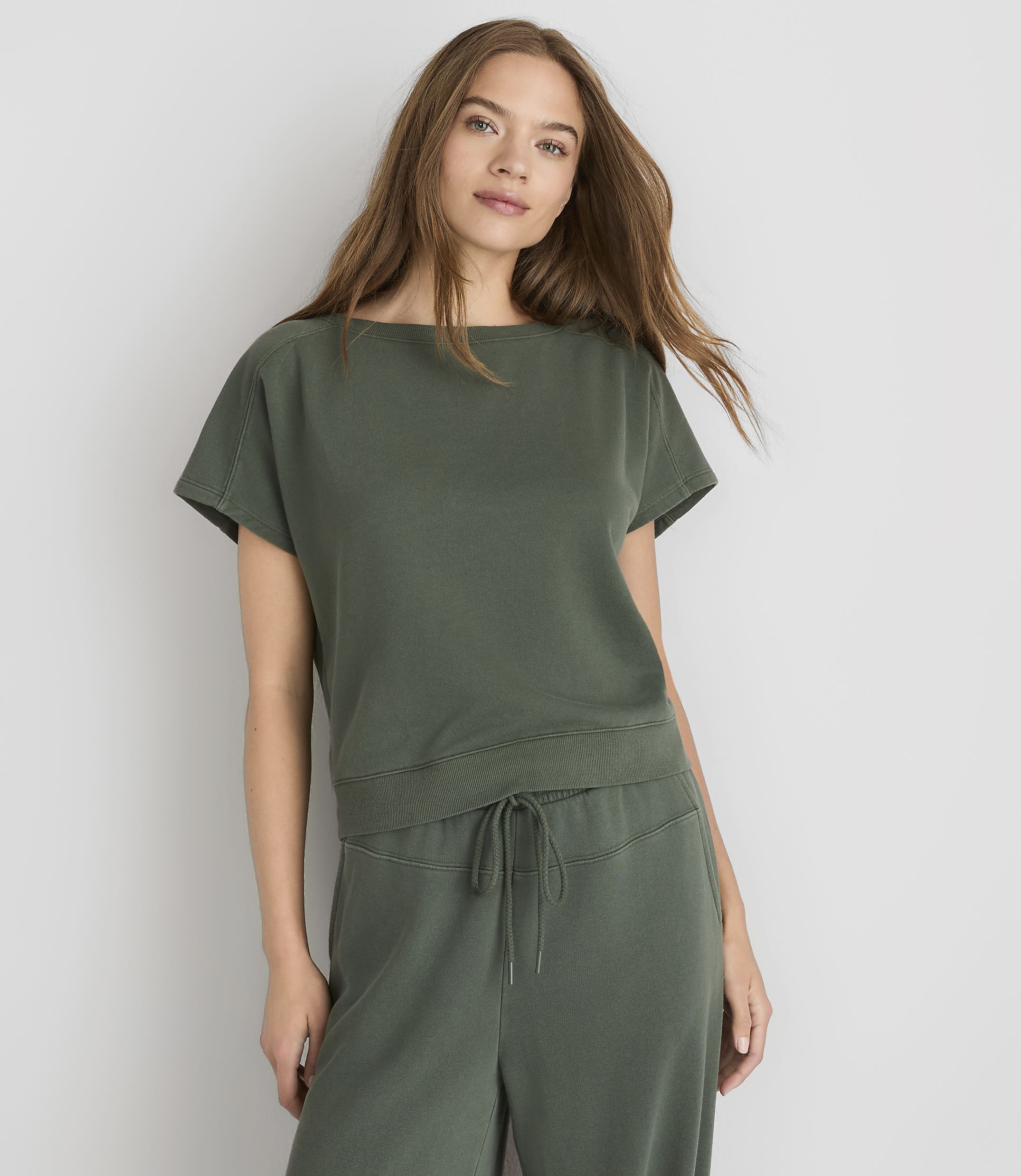 A young woman in a green top and pants stands against a plain gray background.