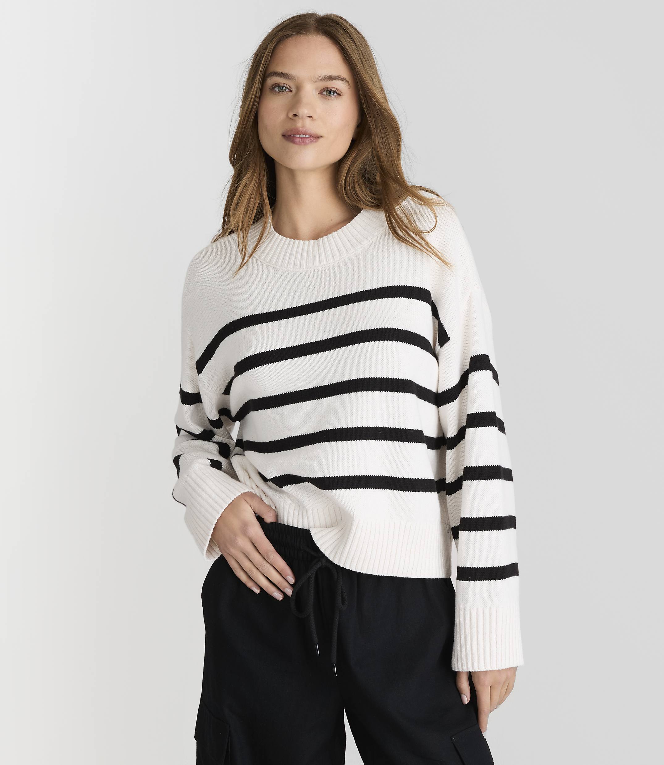 A woman in a striped sweater and black pants stands against a plain white background.