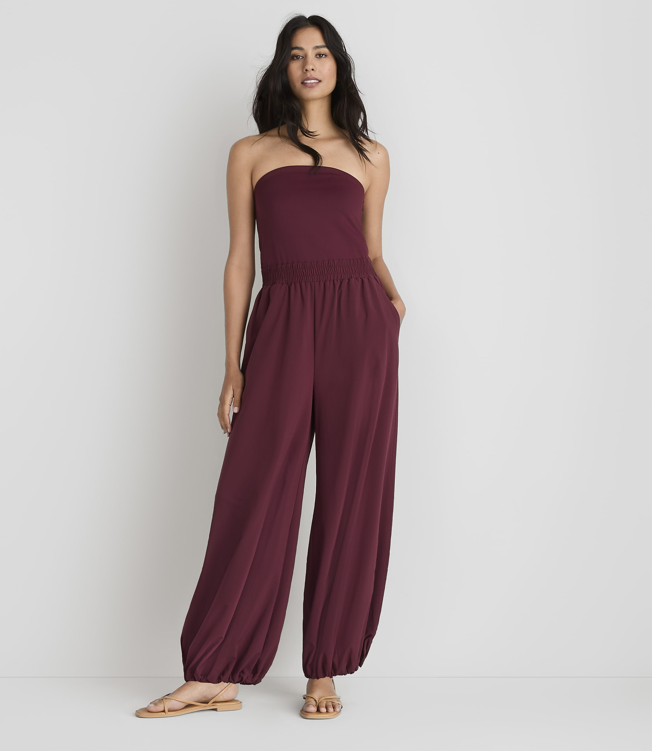A woman in a maroon strapless jumpsuit stands against a plain white background.