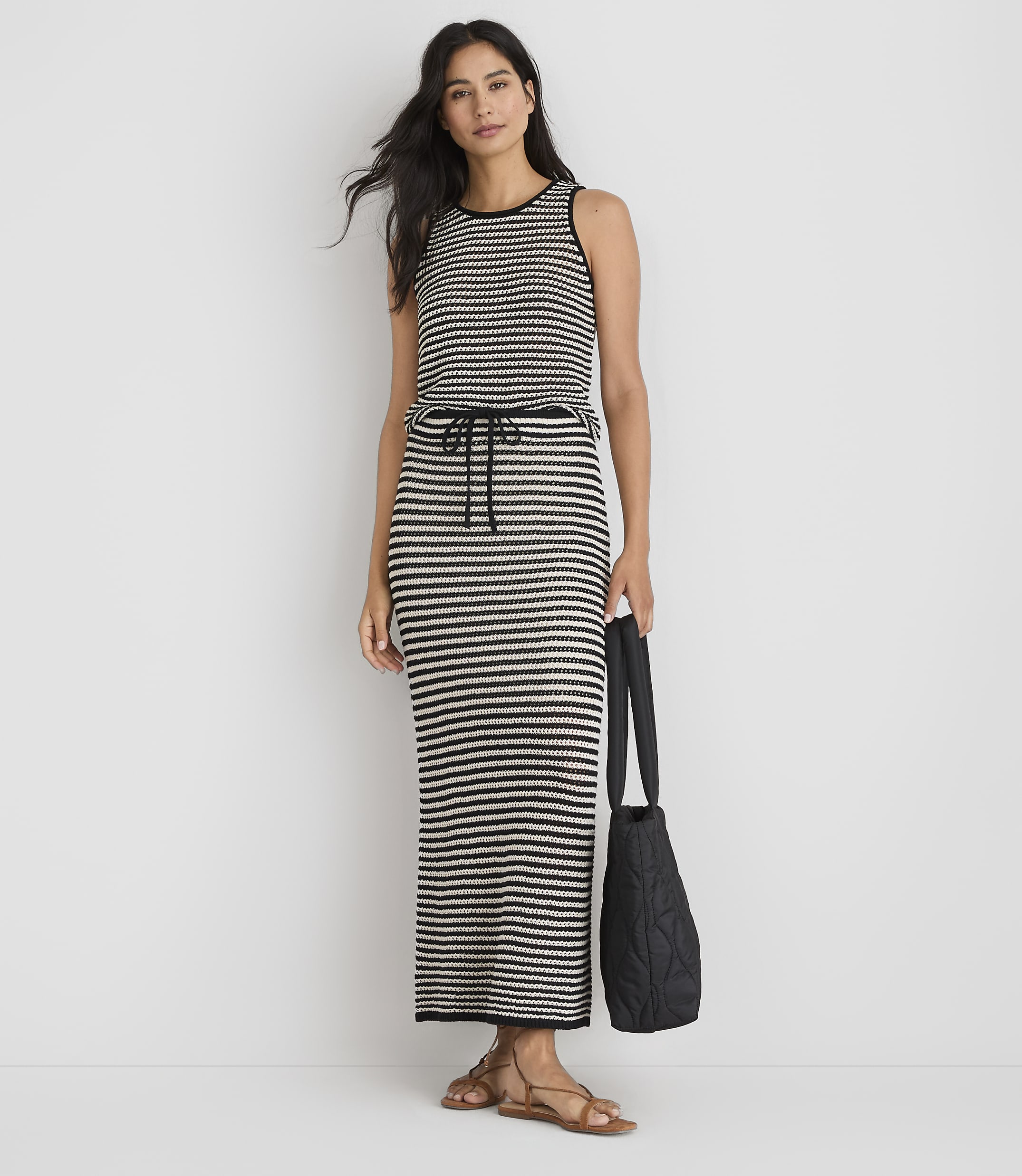 A woman in a striped maxi dress and sandals stands against a plain white background, holding a black quilted tote bag.