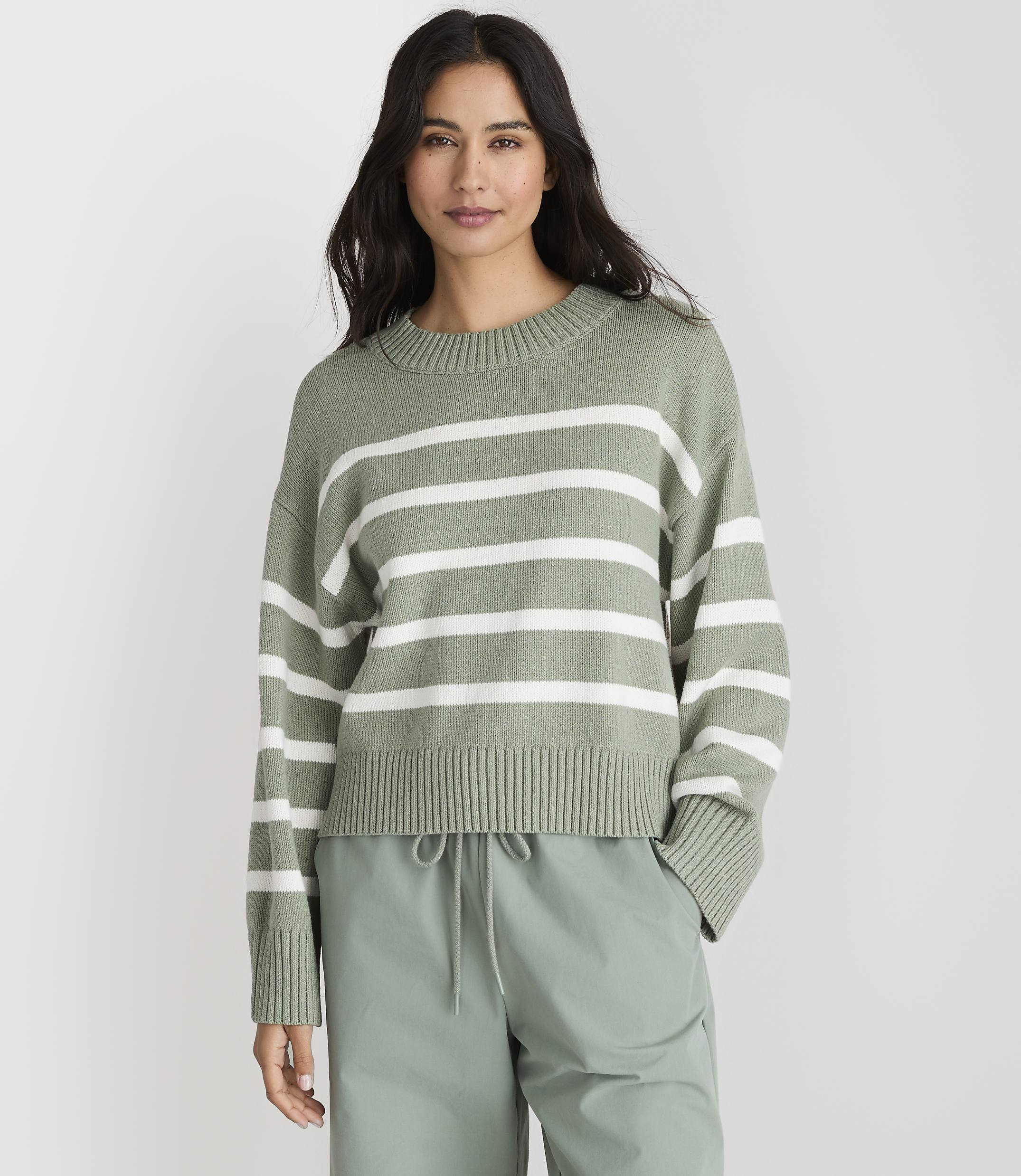 A woman in a green and white striped sweater and light green pants stands against a plain gray background.
