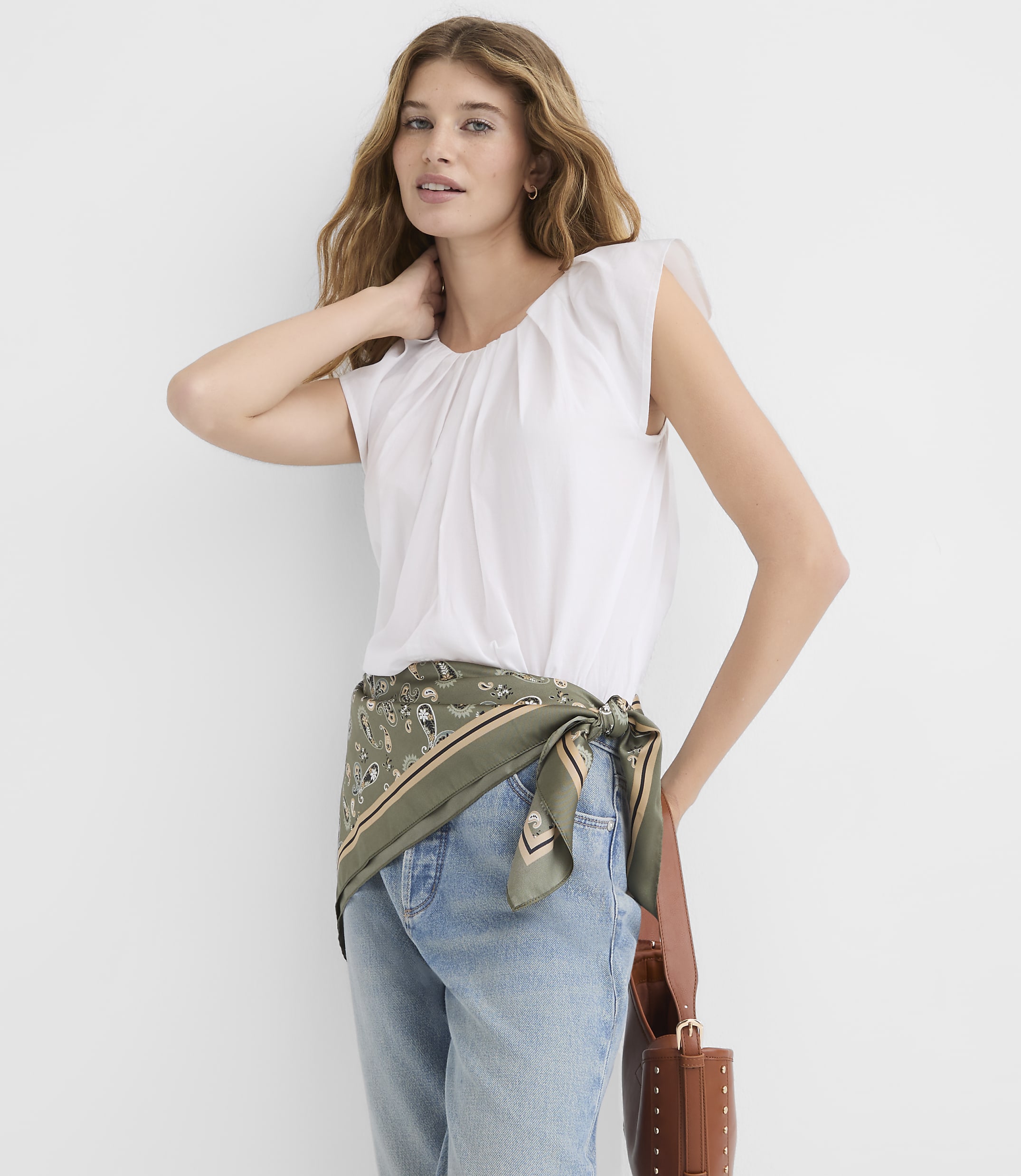 A woman in a white top and jeans, with a green scarf tied around her waist, stands against a plain white background.