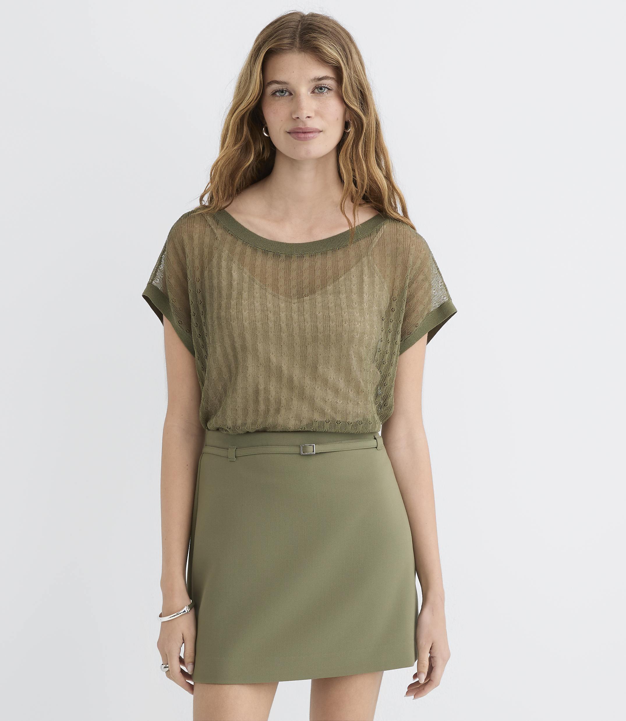 A woman in a sheer olive green top and olive green skirt stands against a plain white background.