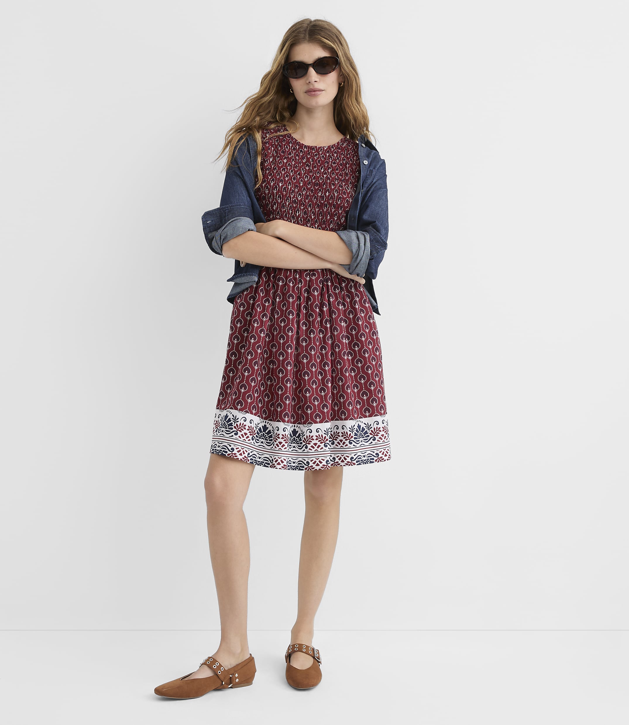 A woman in a patterned dress and denim jacket stands with her arms crossed against a plain white background.
