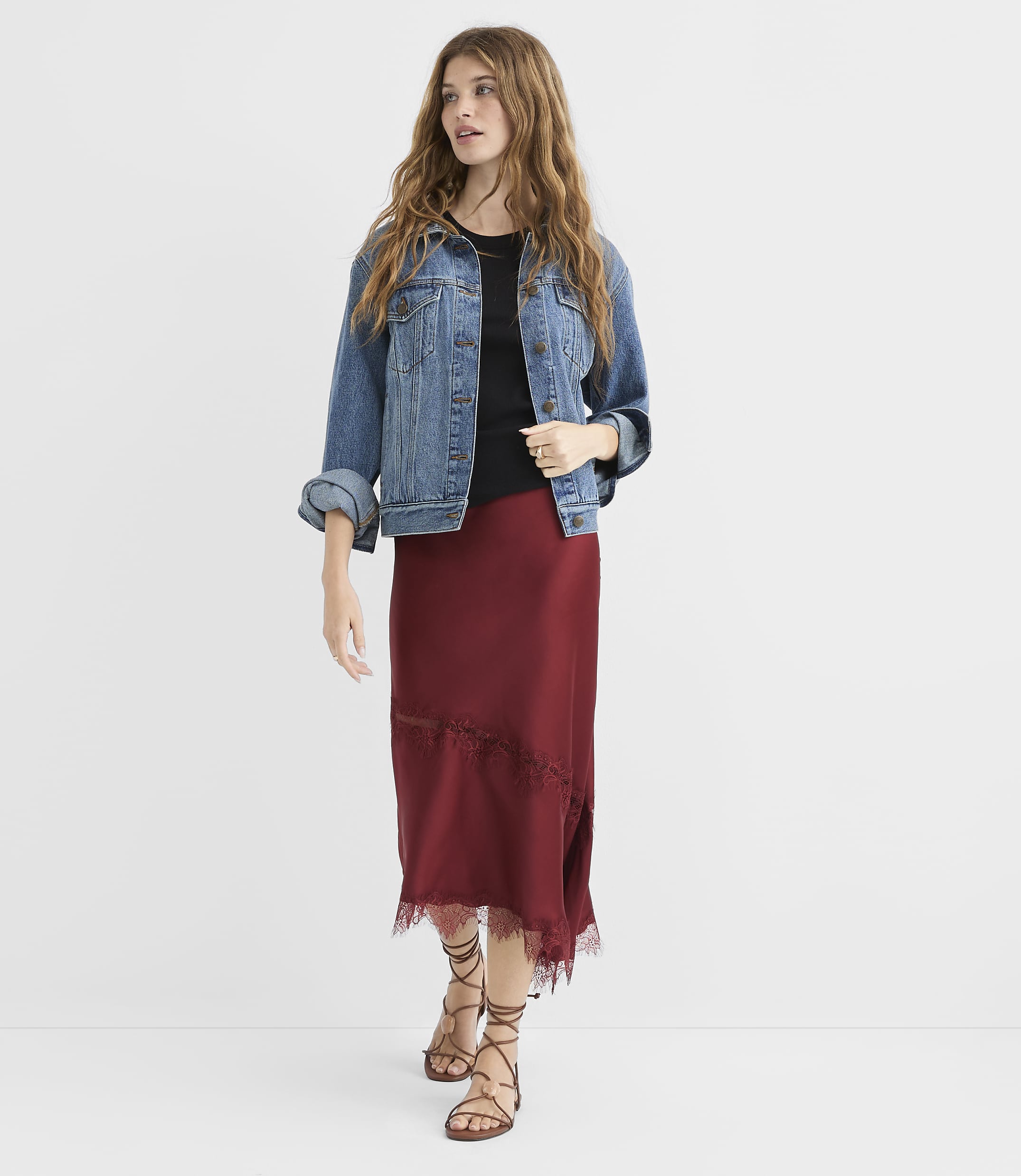A woman in a denim jacket and maroon skirt stands against a plain white background.
