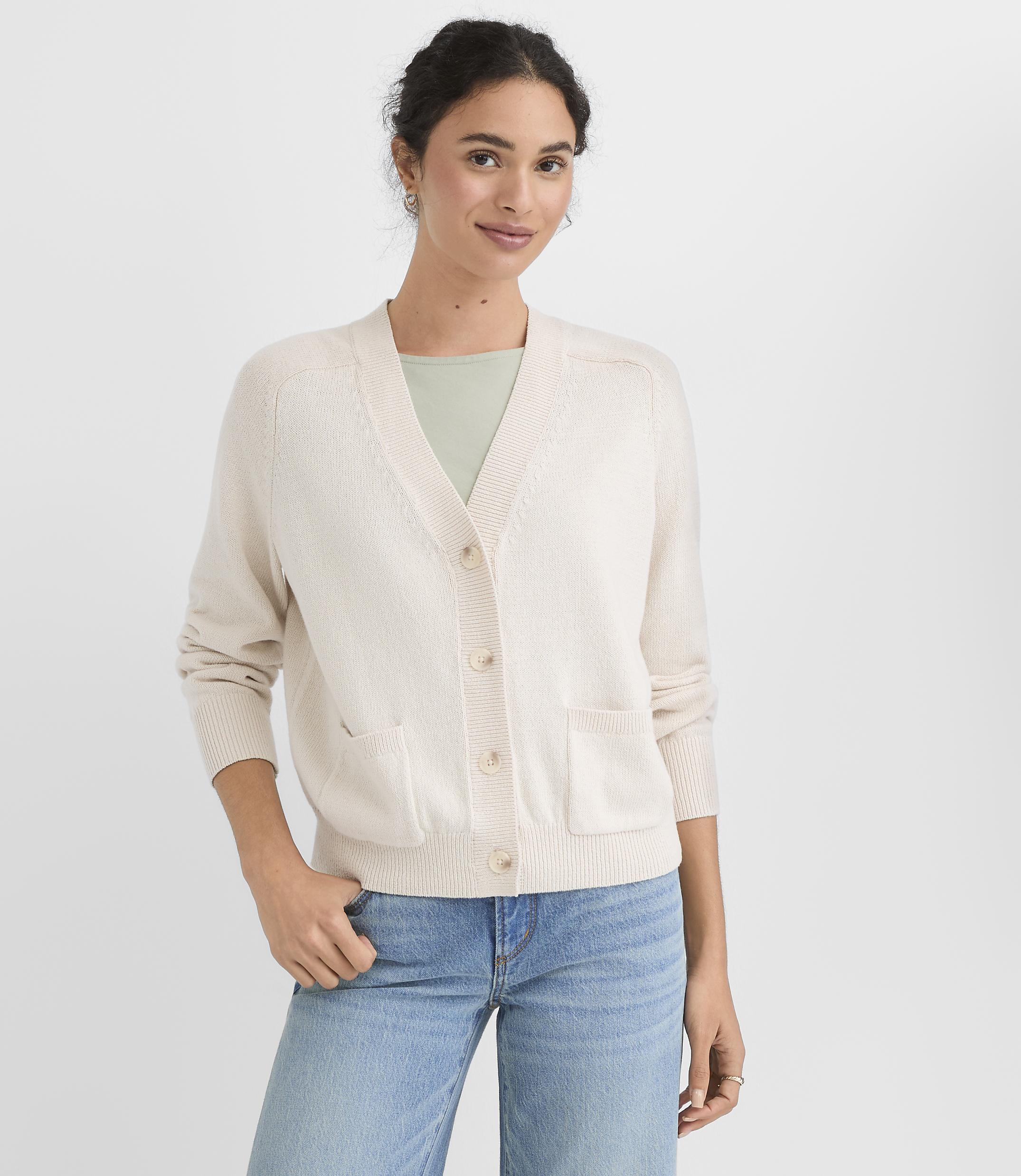 A woman in a cream cardigan and blue jeans stands against a plain white background.