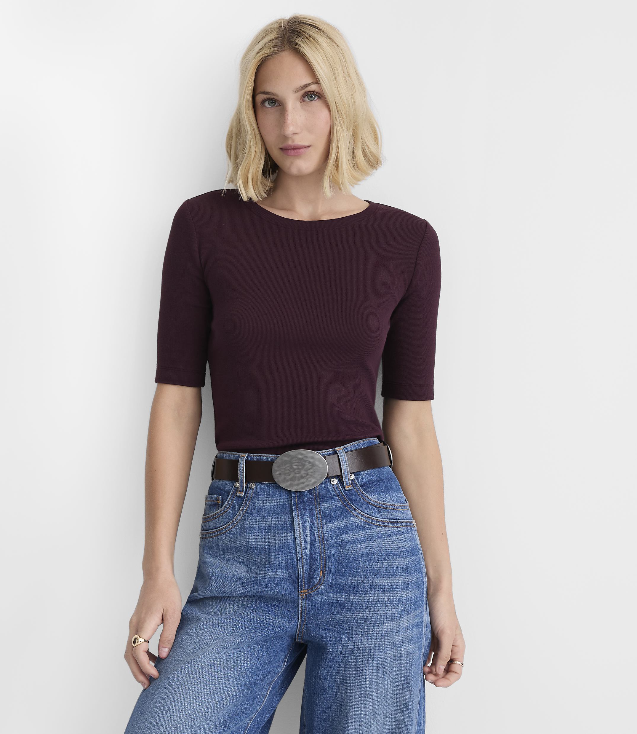 A woman in a maroon ribbed shirt and blue jeans stands against a plain white background.