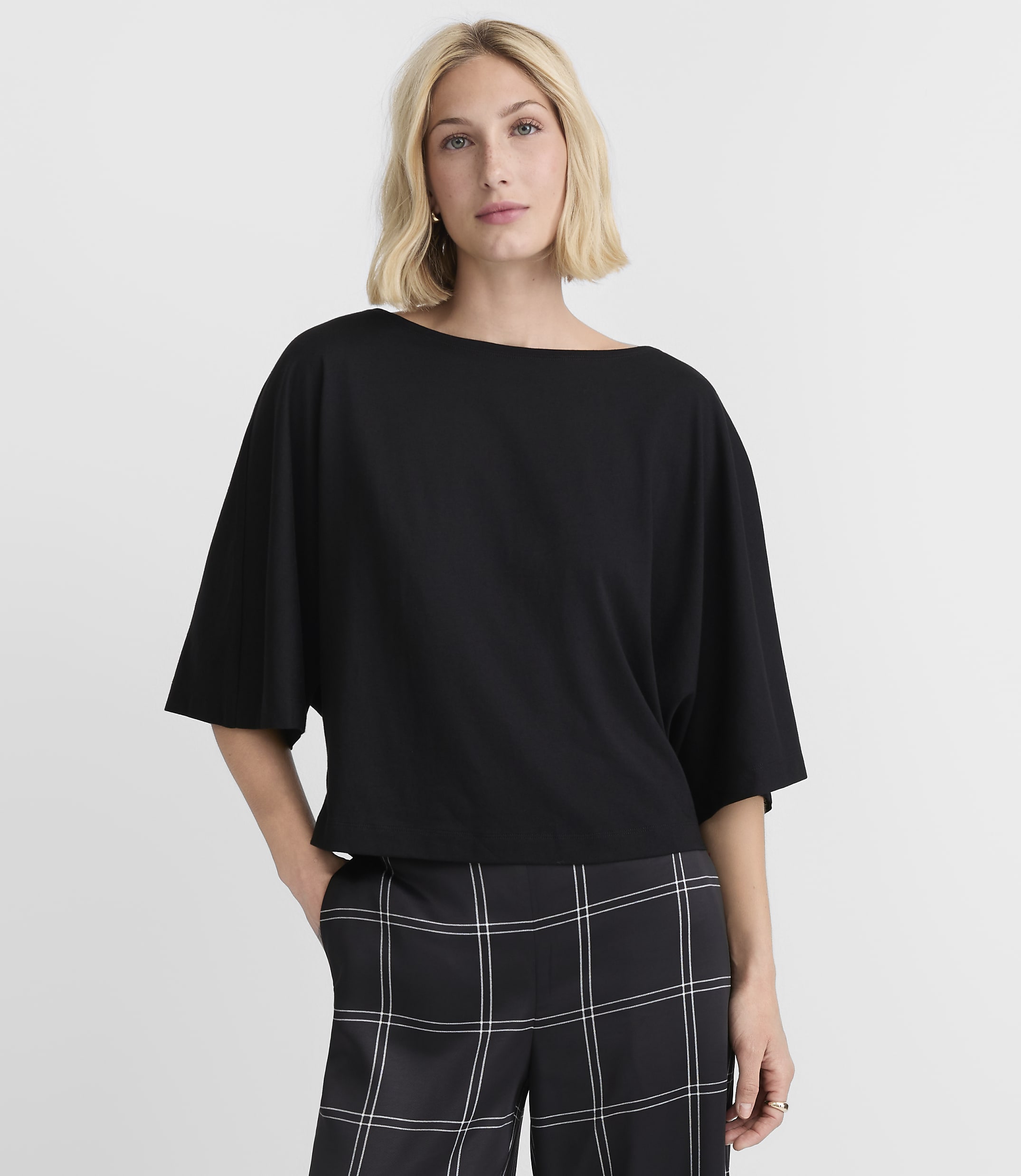 A woman in a black boatneck top and plaid pants stands against a plain white background.