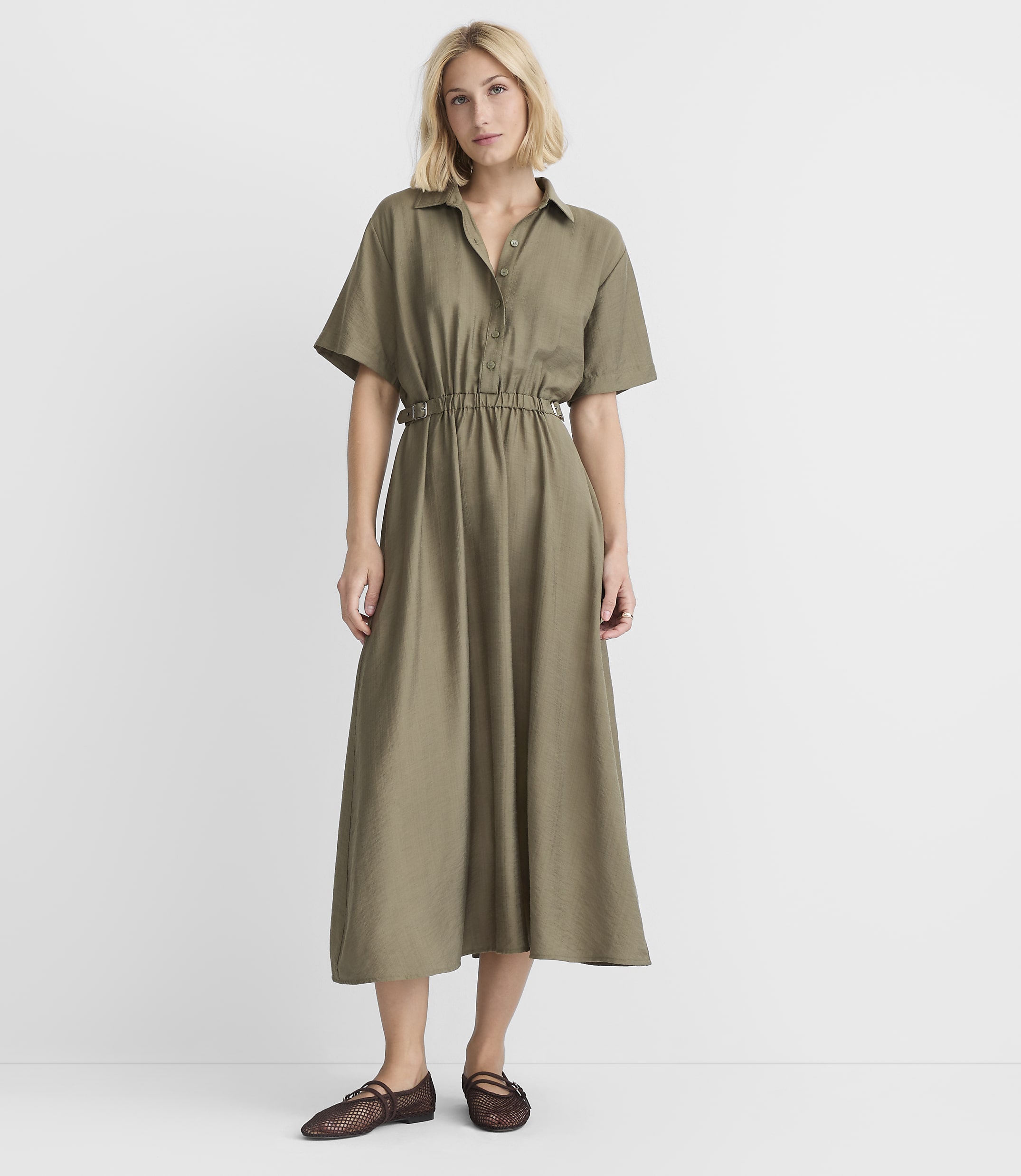 A woman in a green midi dress and brown shoes stands against a plain white background.