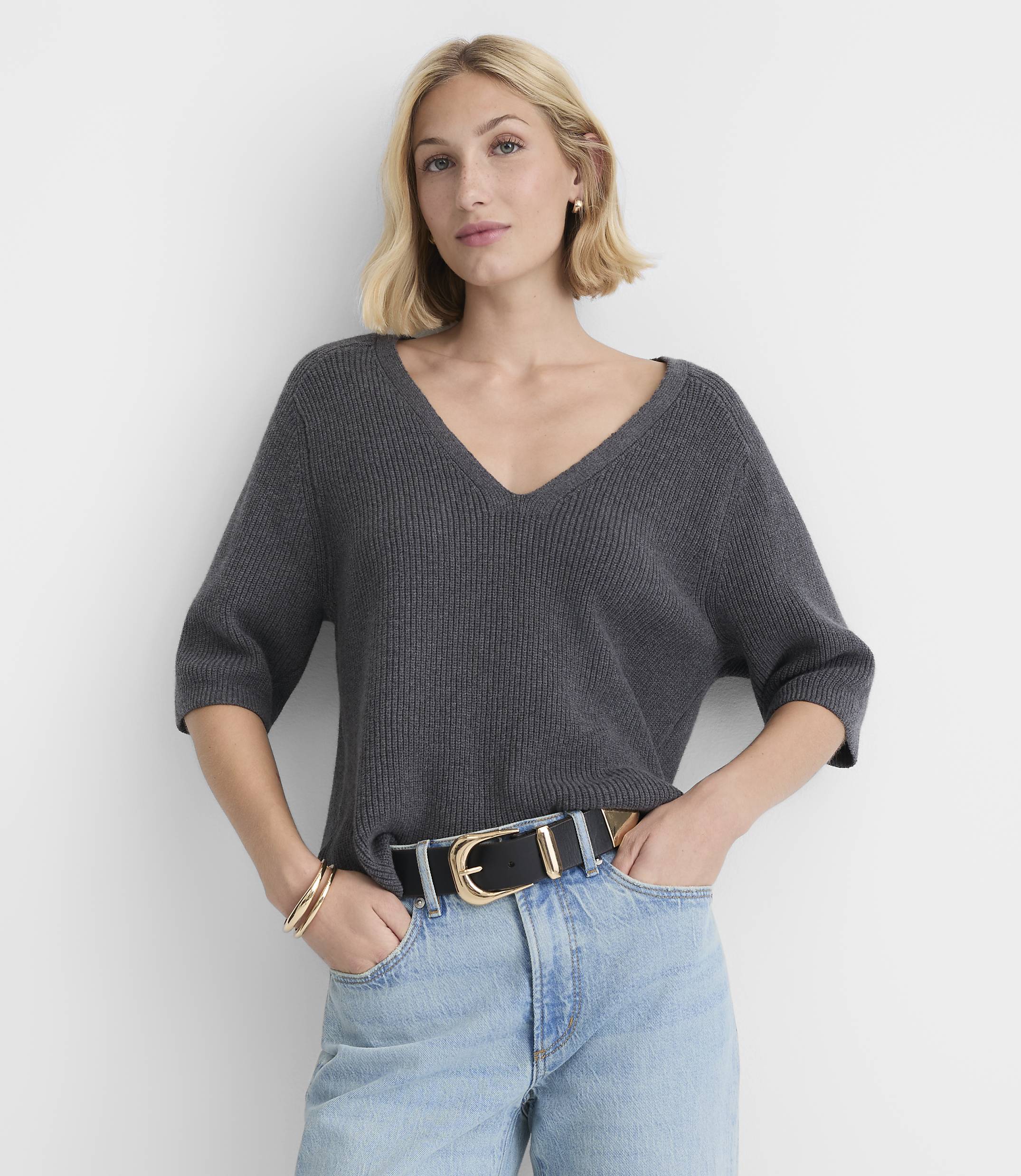 A woman in a dark gray v-neck sweater and light blue jeans stands against a plain white background.