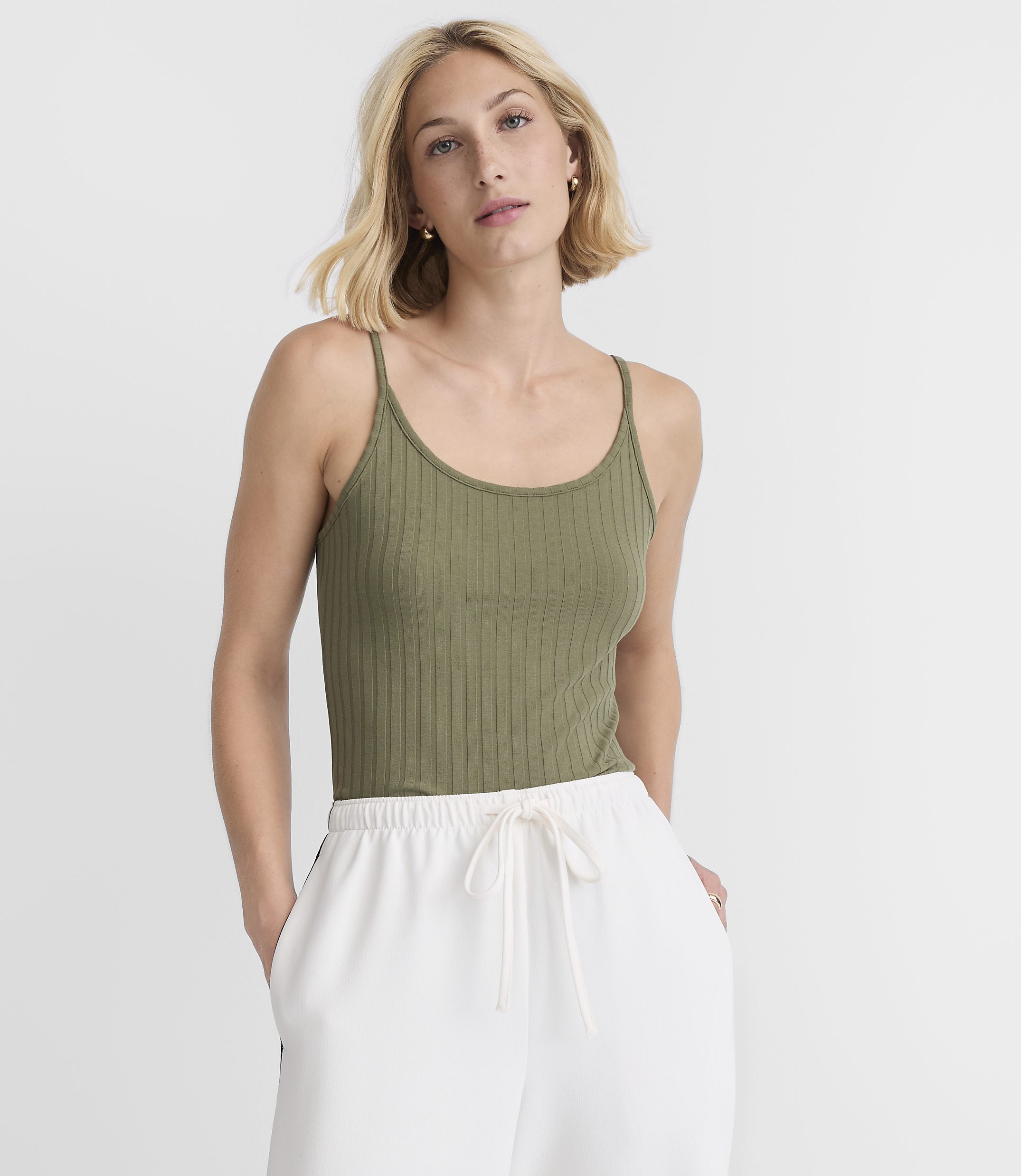 A woman in a green ribbed tank top and white drawstring pants stands against a plain white background.