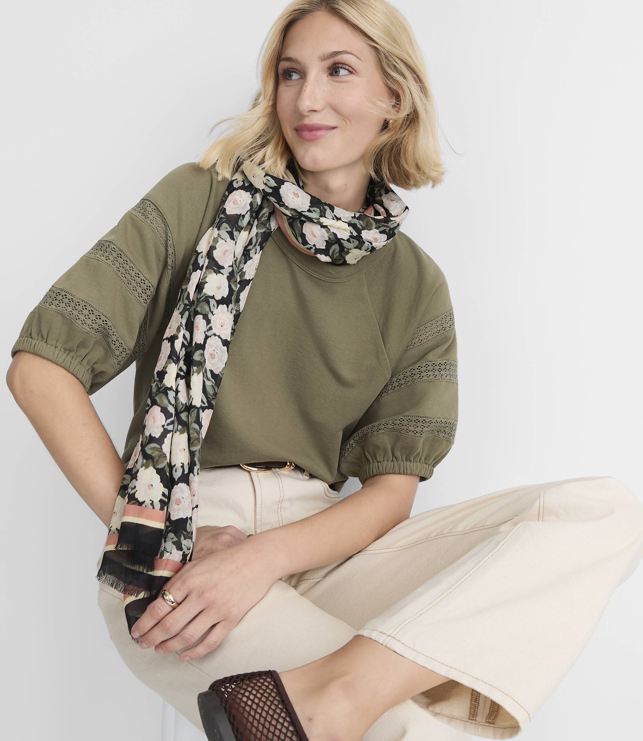 A blonde woman in a green top and cream pants, adorned with a floral scarf, sits against a plain white background.