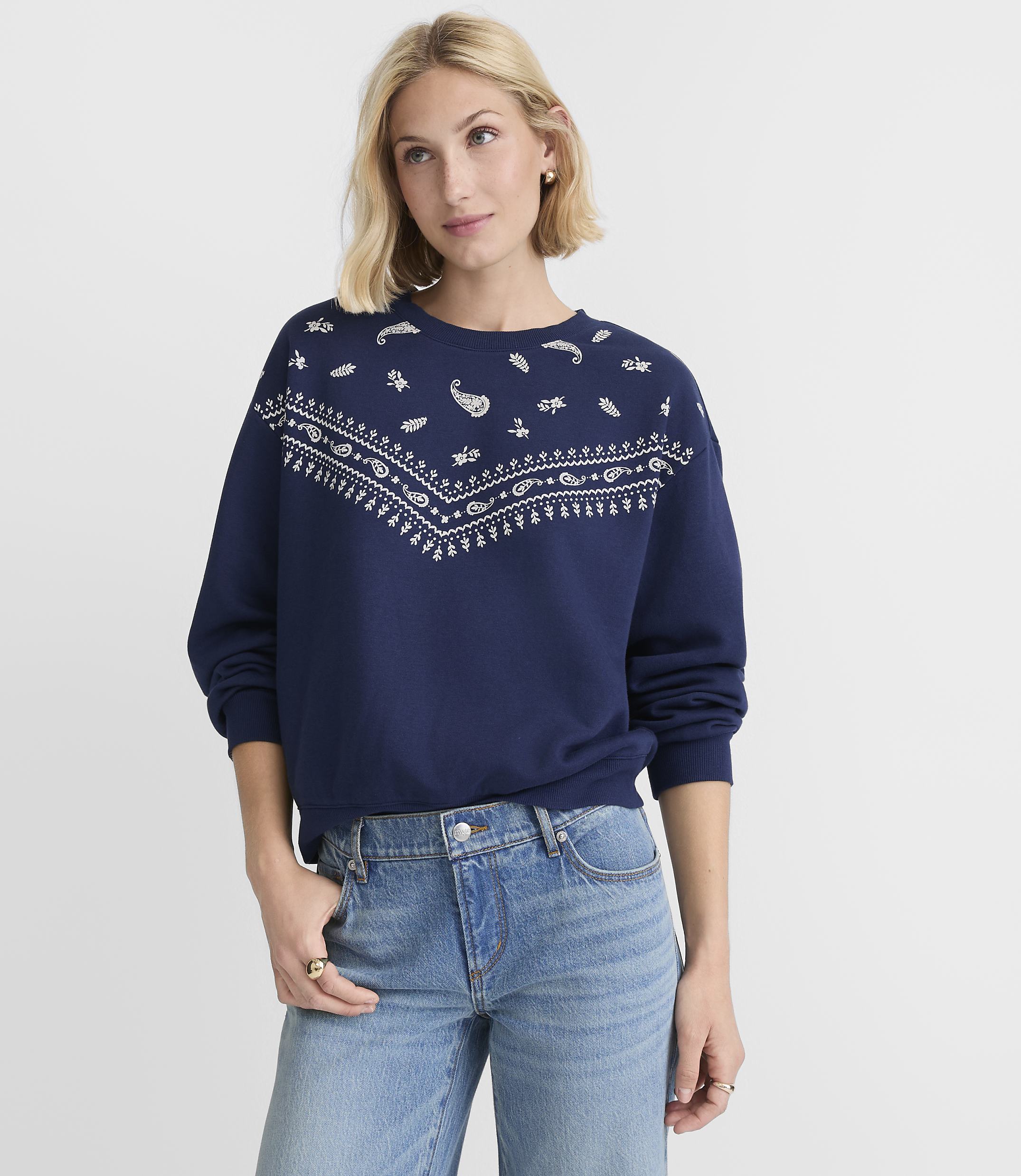 A woman in a navy sweatshirt with a white paisley pattern and blue jeans stands against a plain white background.
