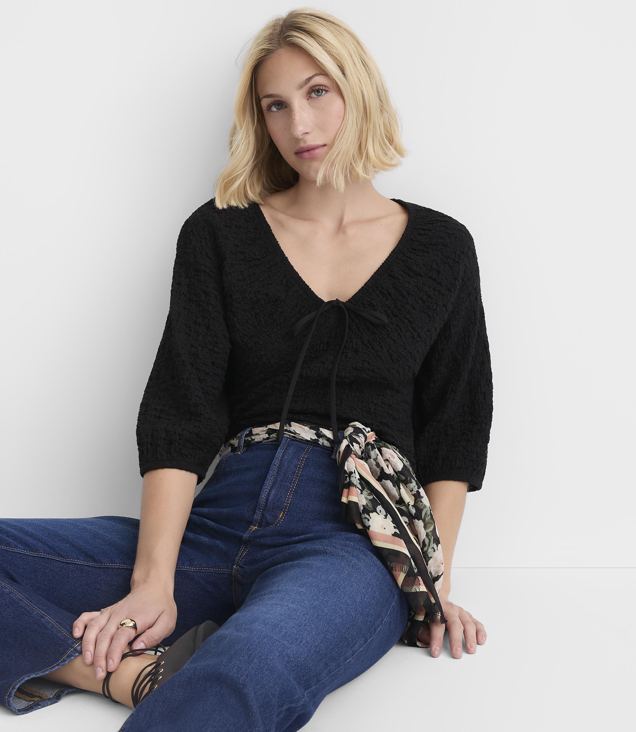 A woman in a black v-neck top and blue jeans sits against a plain white background.