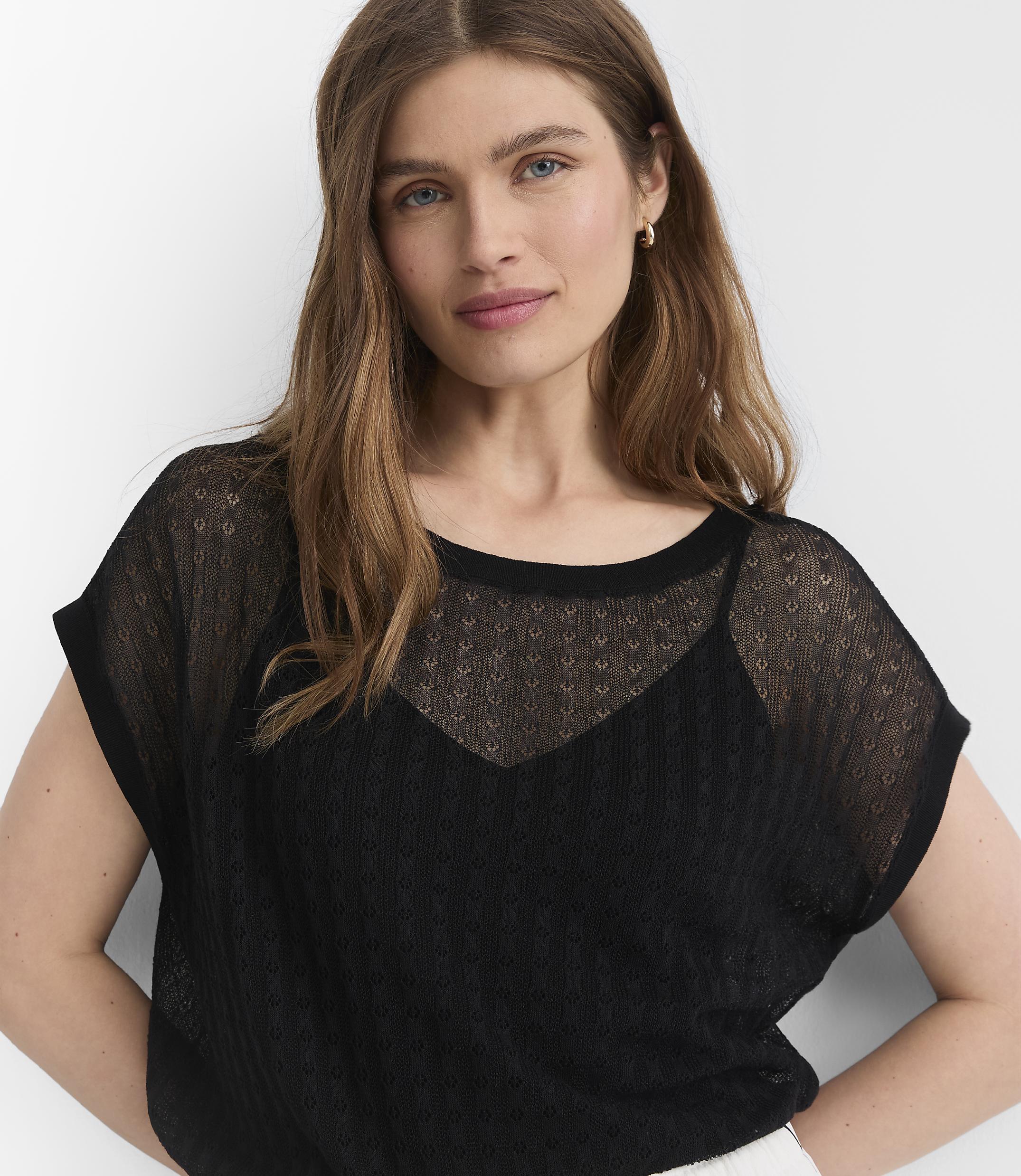A woman with long brown hair wears a sheer black top over a camisole, with a plain white background.
