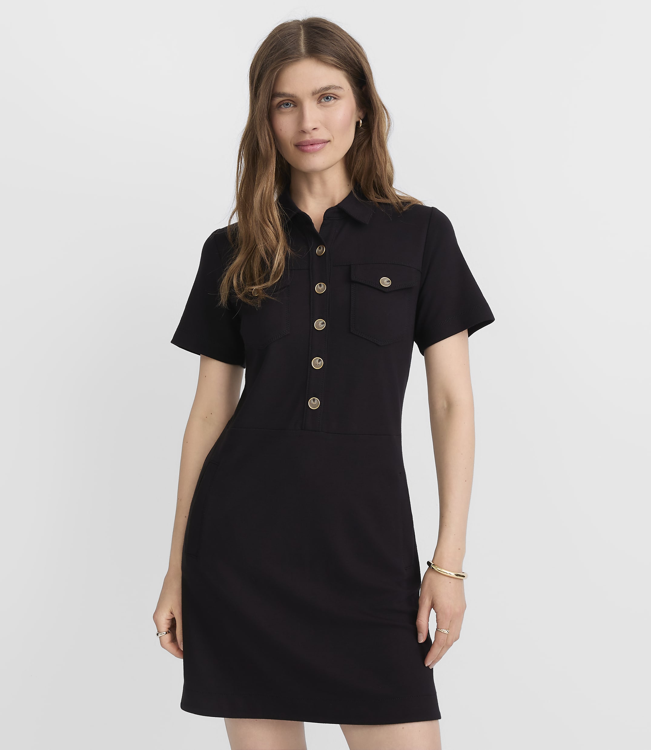 A woman in a black collared dress with gold buttons stands against a plain gray background.