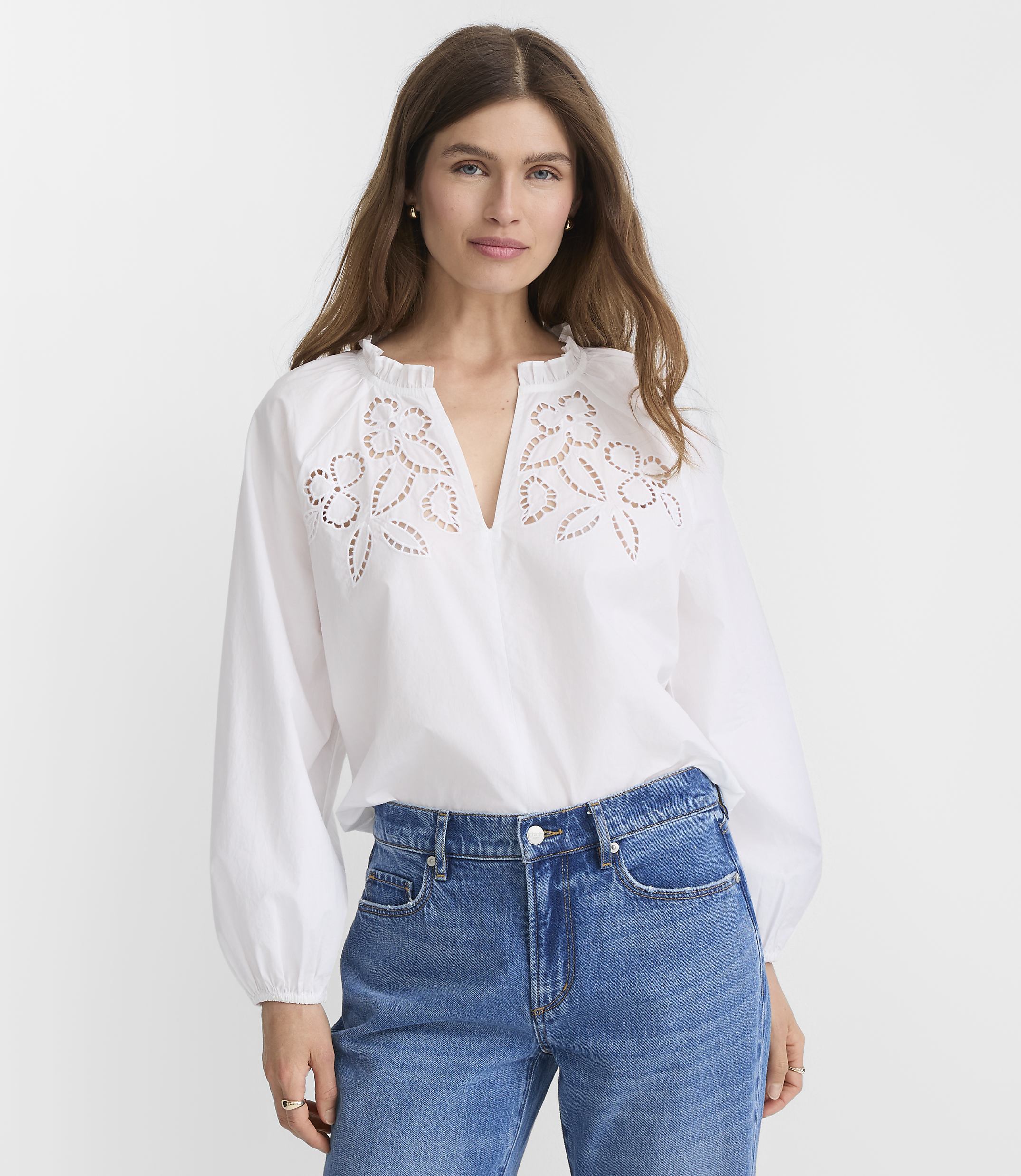 A woman in a white embroidered blouse and blue jeans stands against a plain white background.