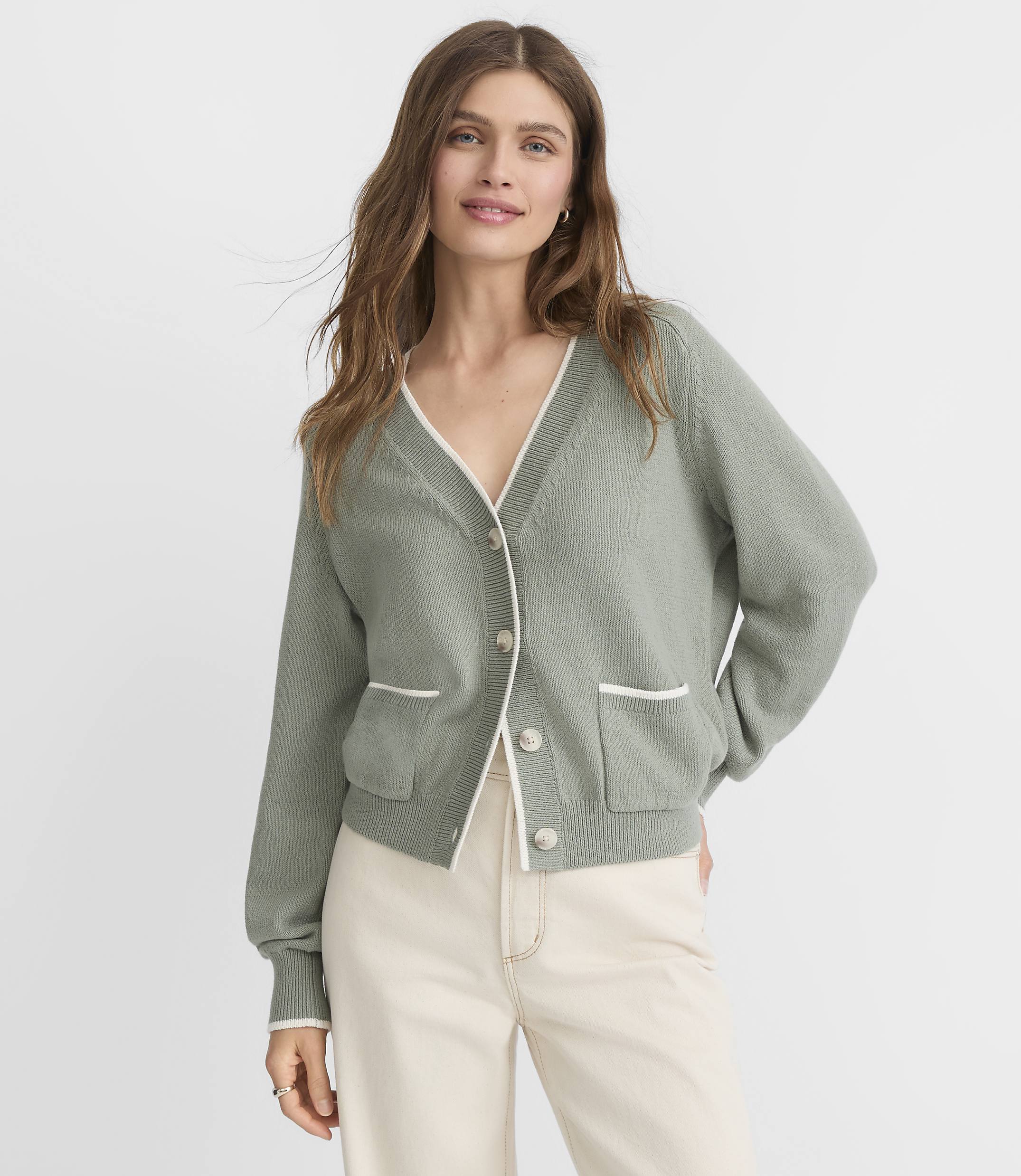 A woman in a sage green cardigan with white trim stands against a plain, light gray background.