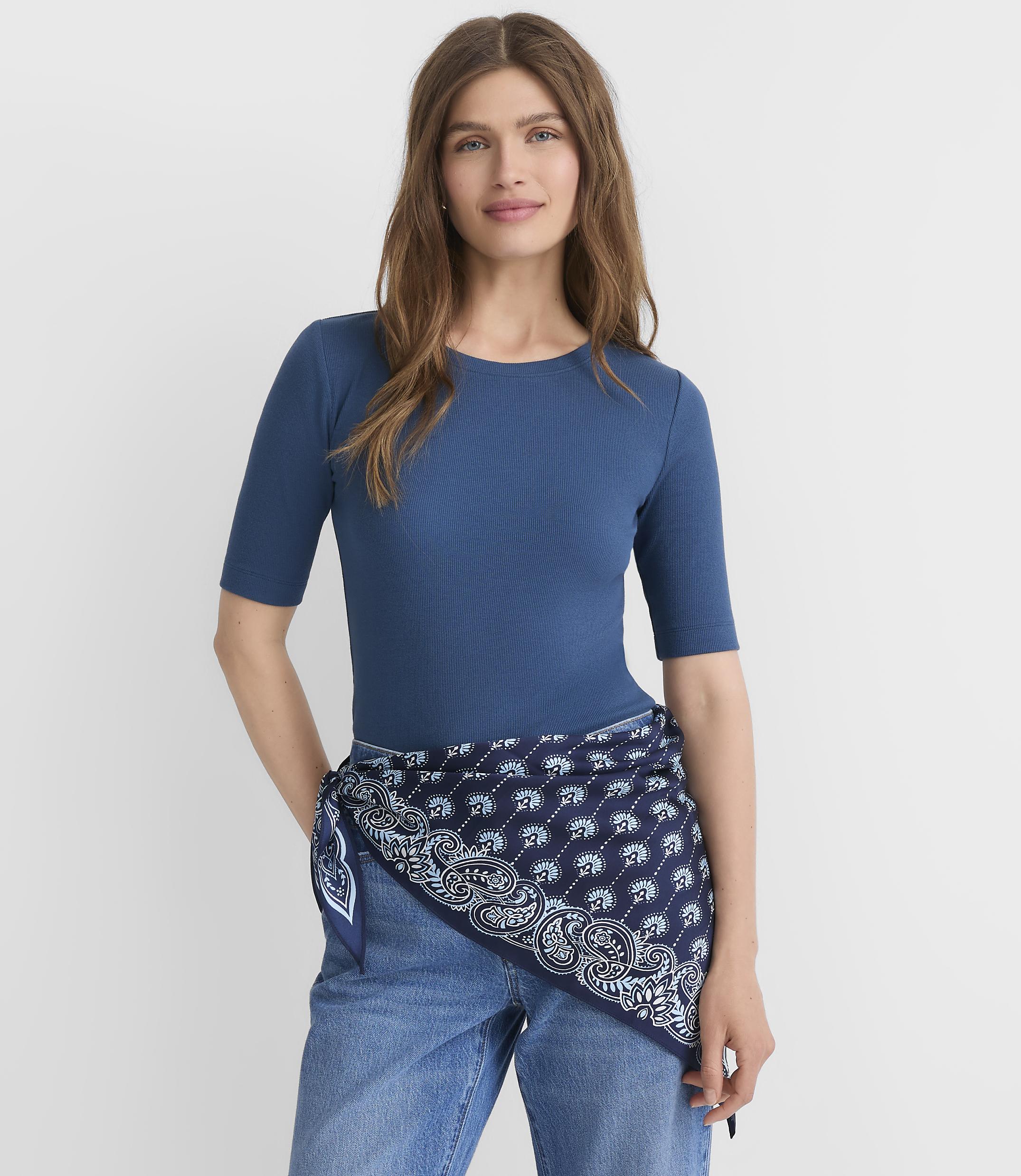 A woman in a blue top and jeans stands against a plain white background, with a patterned scarf tied around her waist.