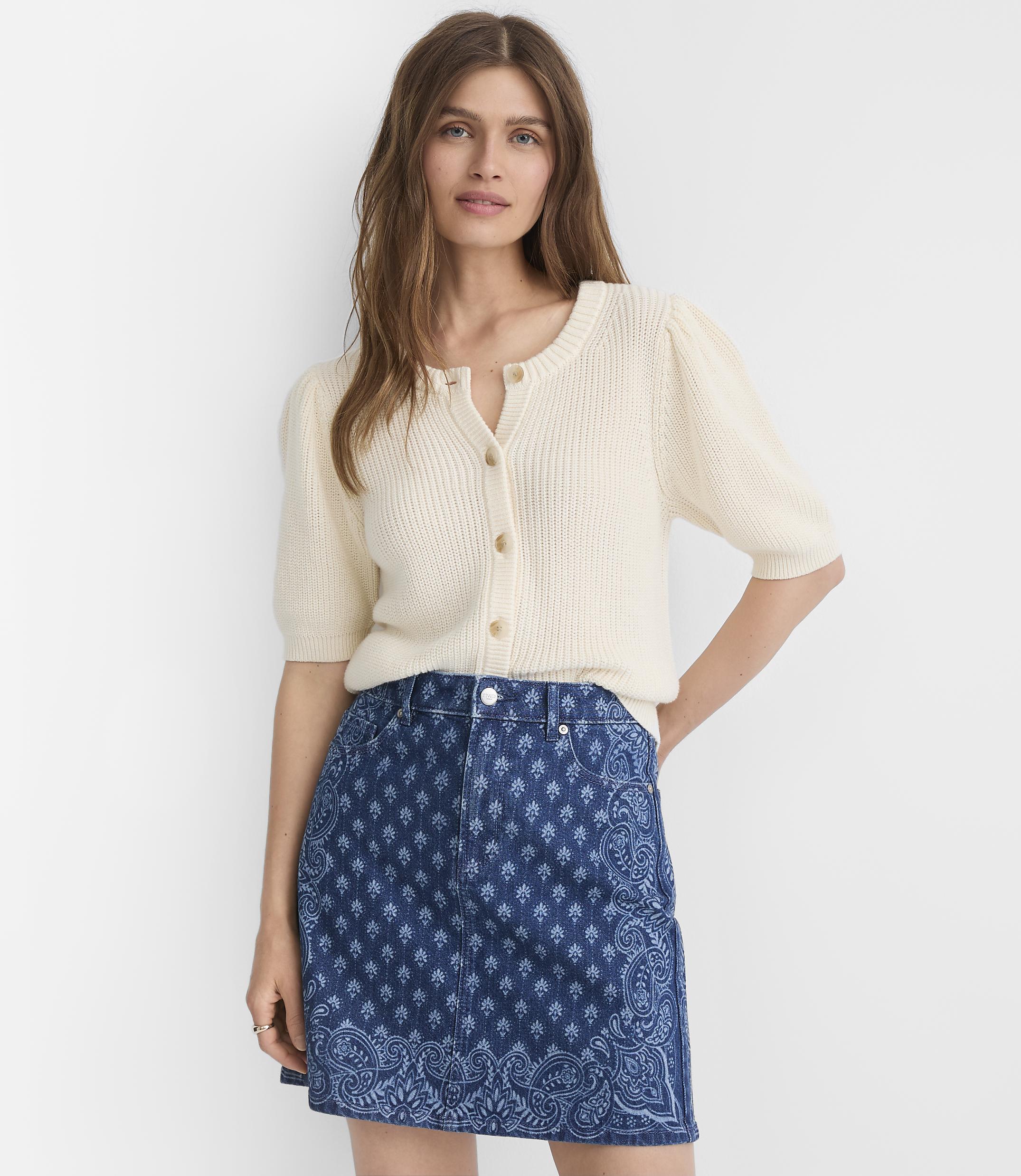 A woman in a cream-colored cardigan and a patterned denim skirt stands against a plain white background.