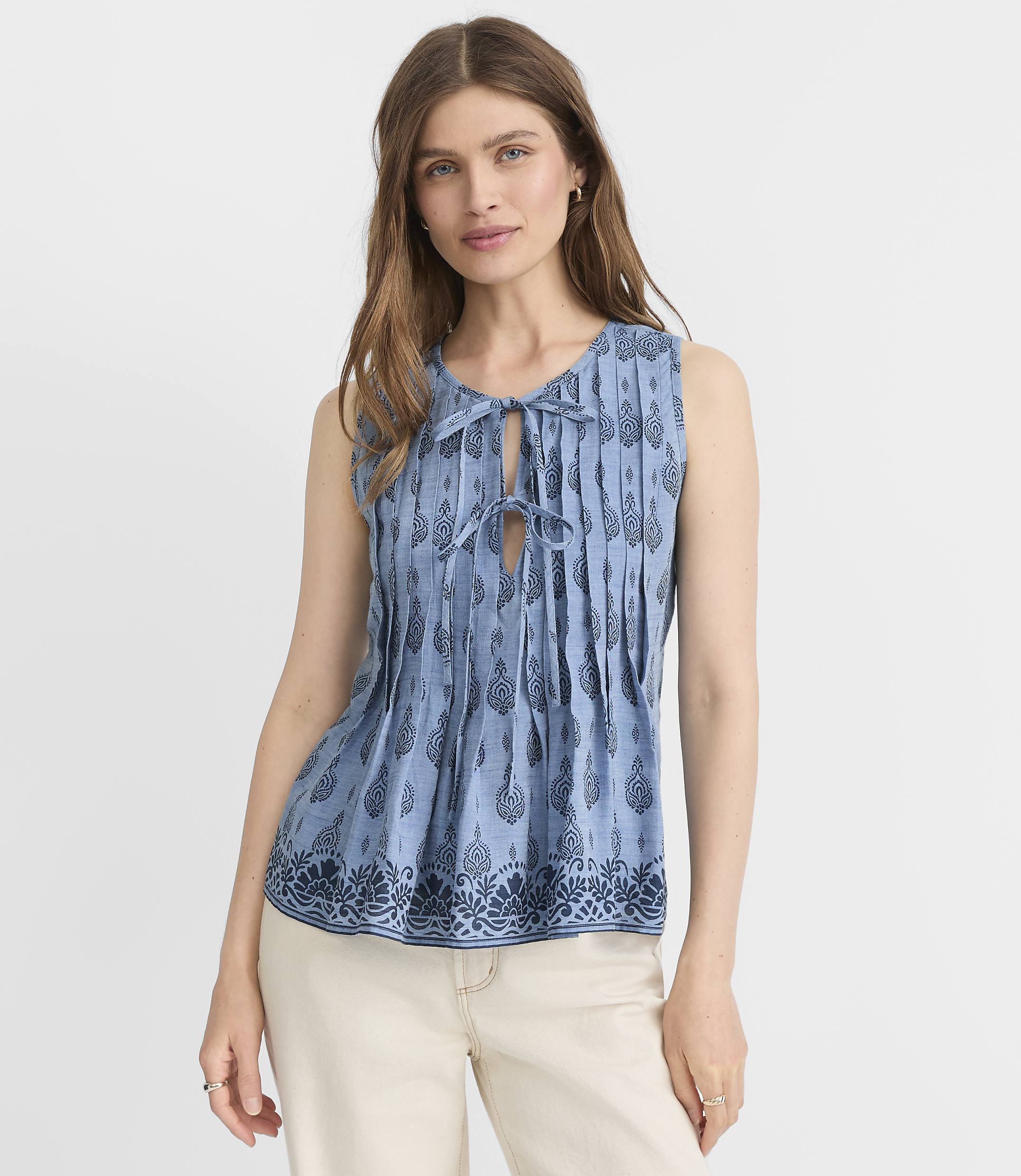 A woman in a blue patterned sleeveless top and cream pants stands against a plain white background.