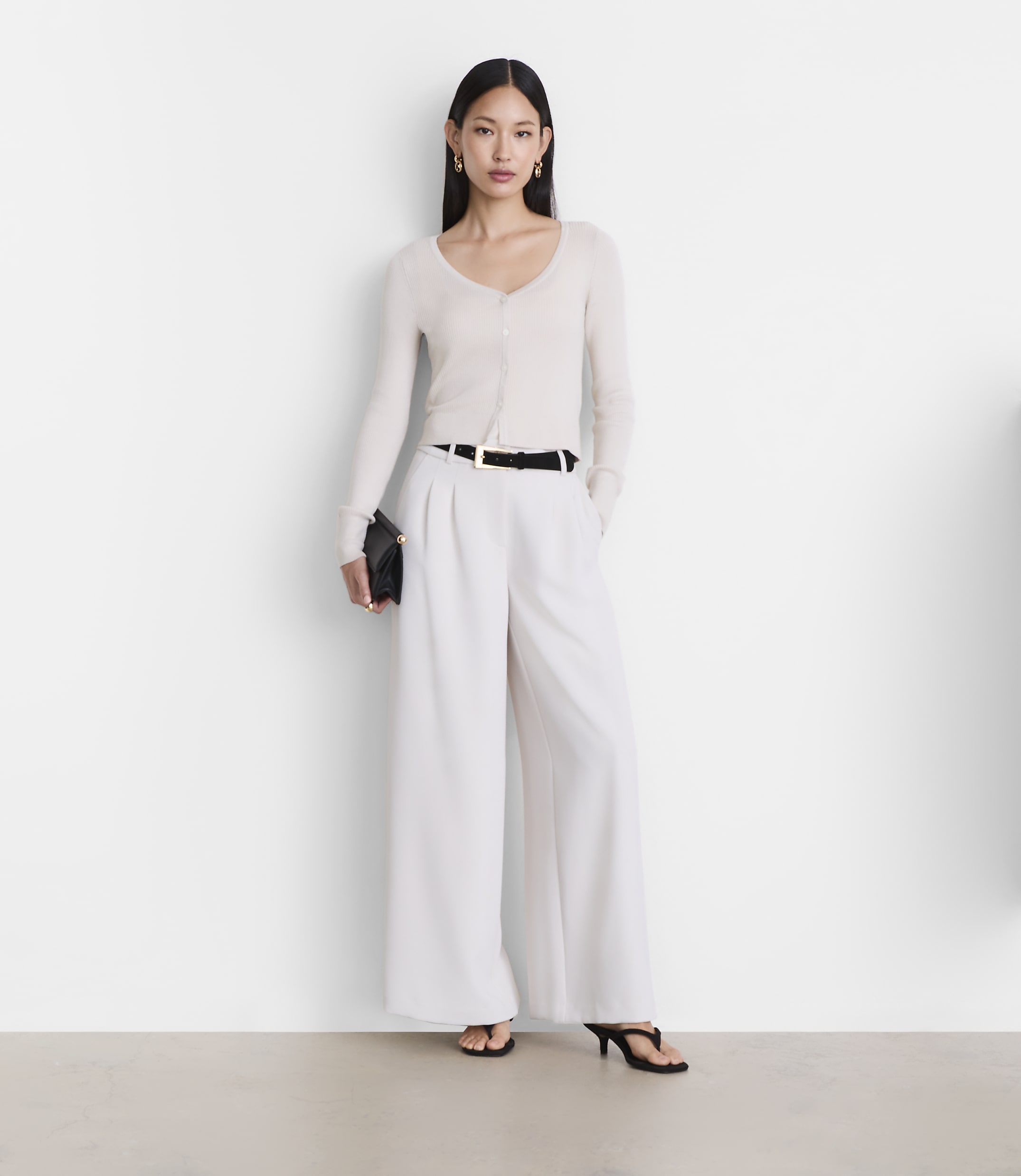 A woman in a cream cardigan and white wide-leg pants stands against a plain white background.
