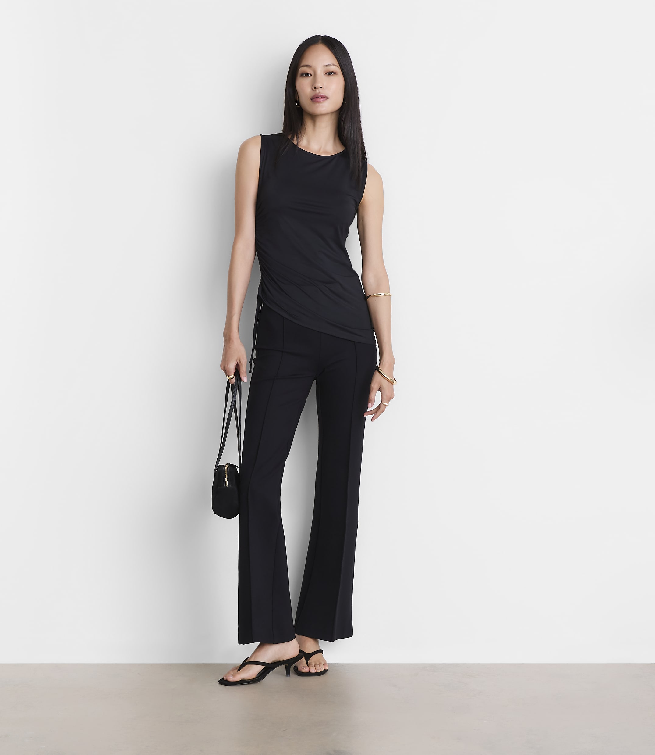 A woman in a black tank top and flared pants stands against a plain white background, holding a small black purse.