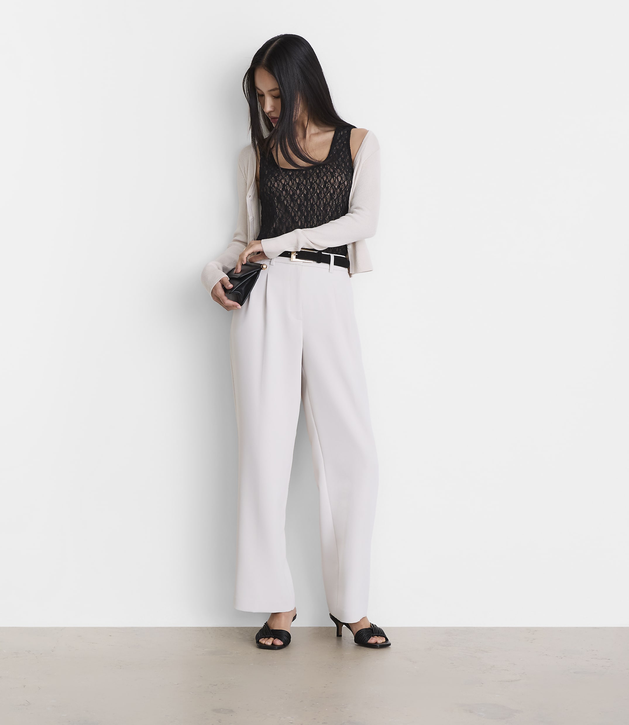 A woman in a black lace top, white pants, and a light cardigan stands against a plain white wall, holding a small black clutch.