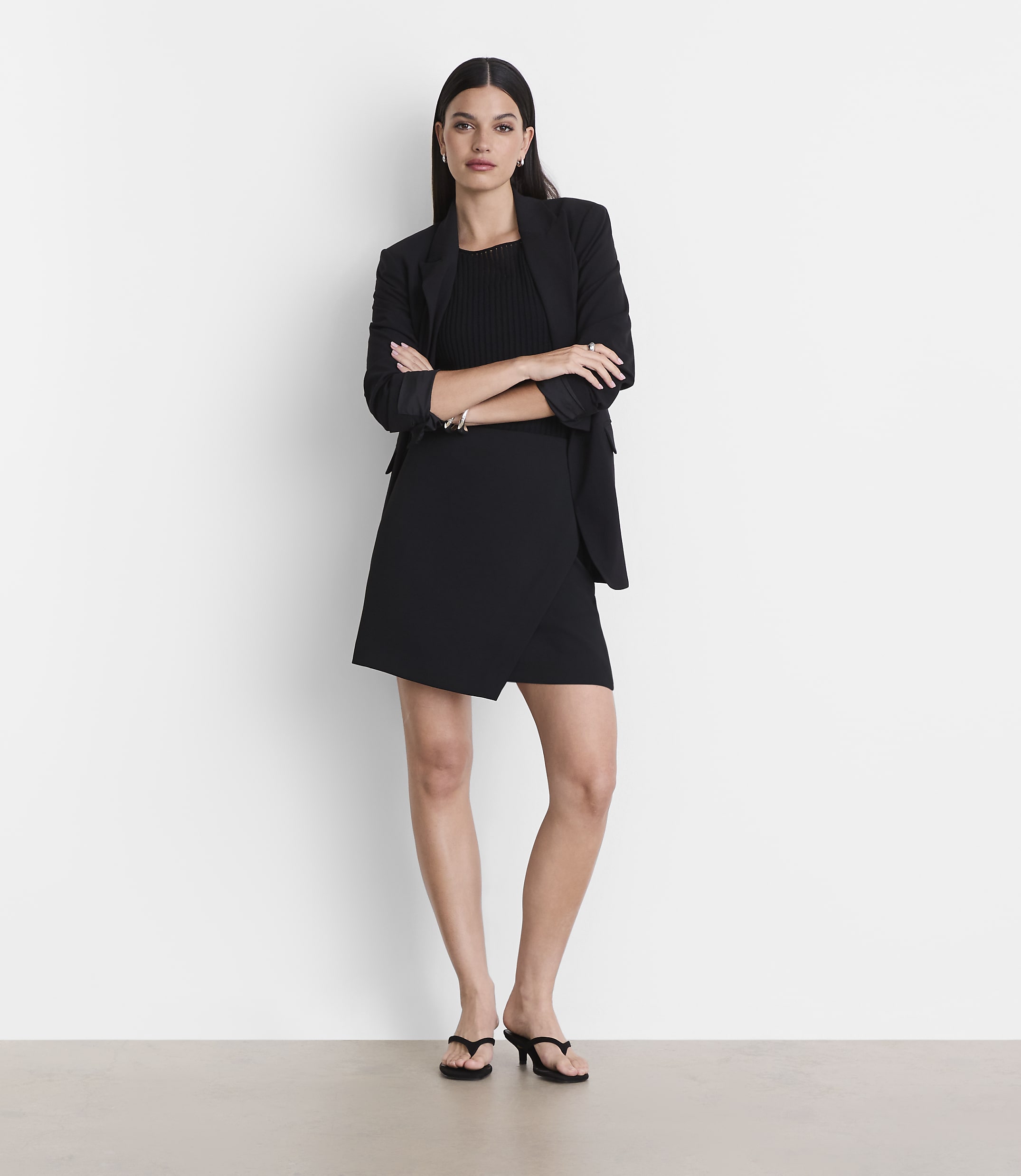 A woman in a black dress and blazer stands with her arms crossed against a plain white background.