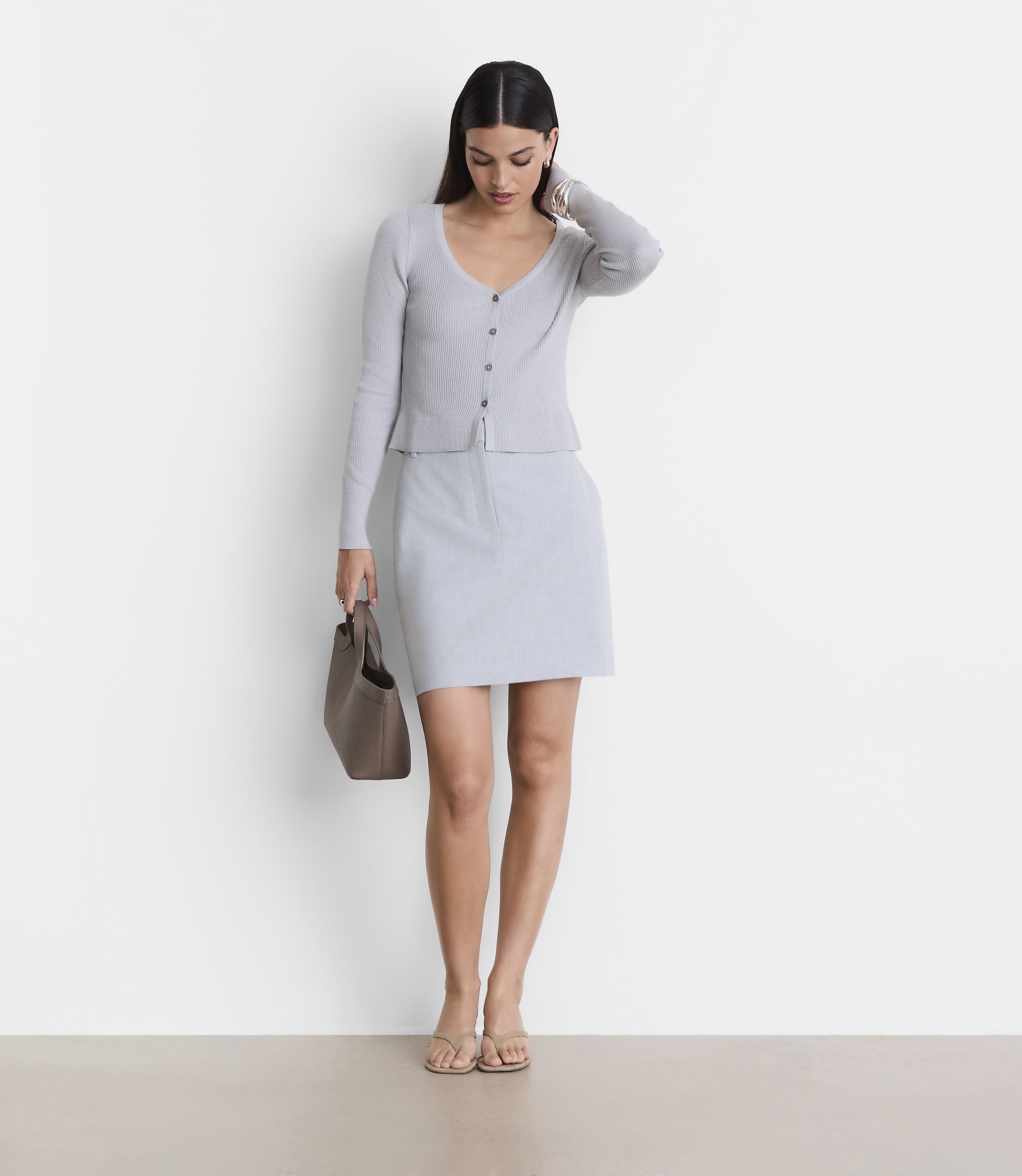 A woman in a light gray cardigan and skirt stands with a brown purse against a plain white background.