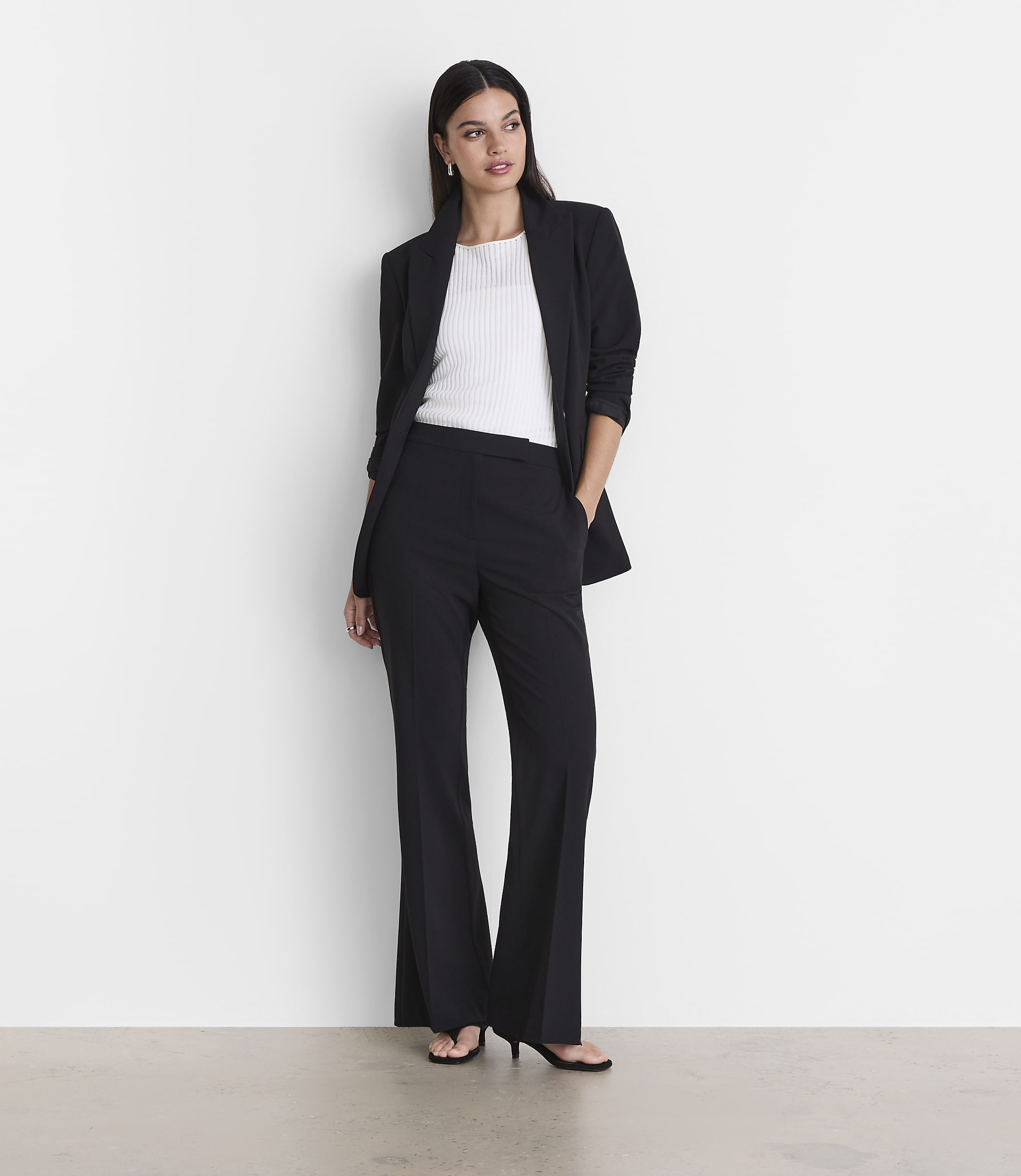 A woman in a black blazer and flared pants stands against a plain white background with a light gray floor in the foreground.