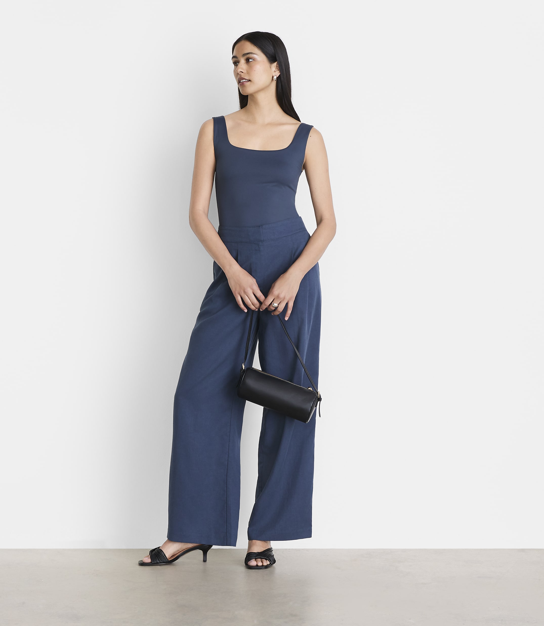 A woman in a blue tank top and wide-leg pants stands against a plain white background, holding a black cylindrical purse.