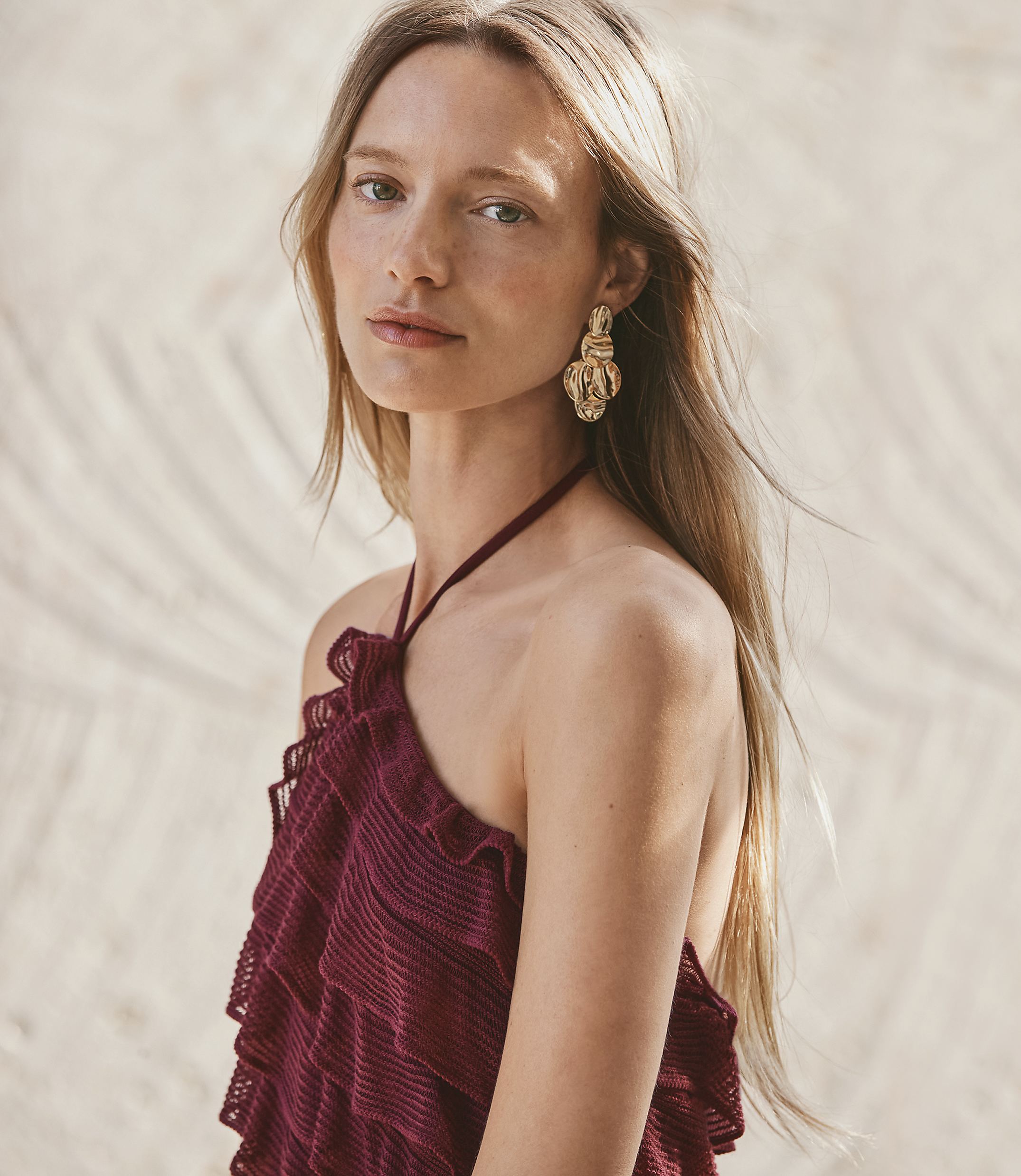 A woman with long blonde hair and large gold earrings wears a ruffled maroon halter top, with a sandy, palm-leaf-patterned background.
