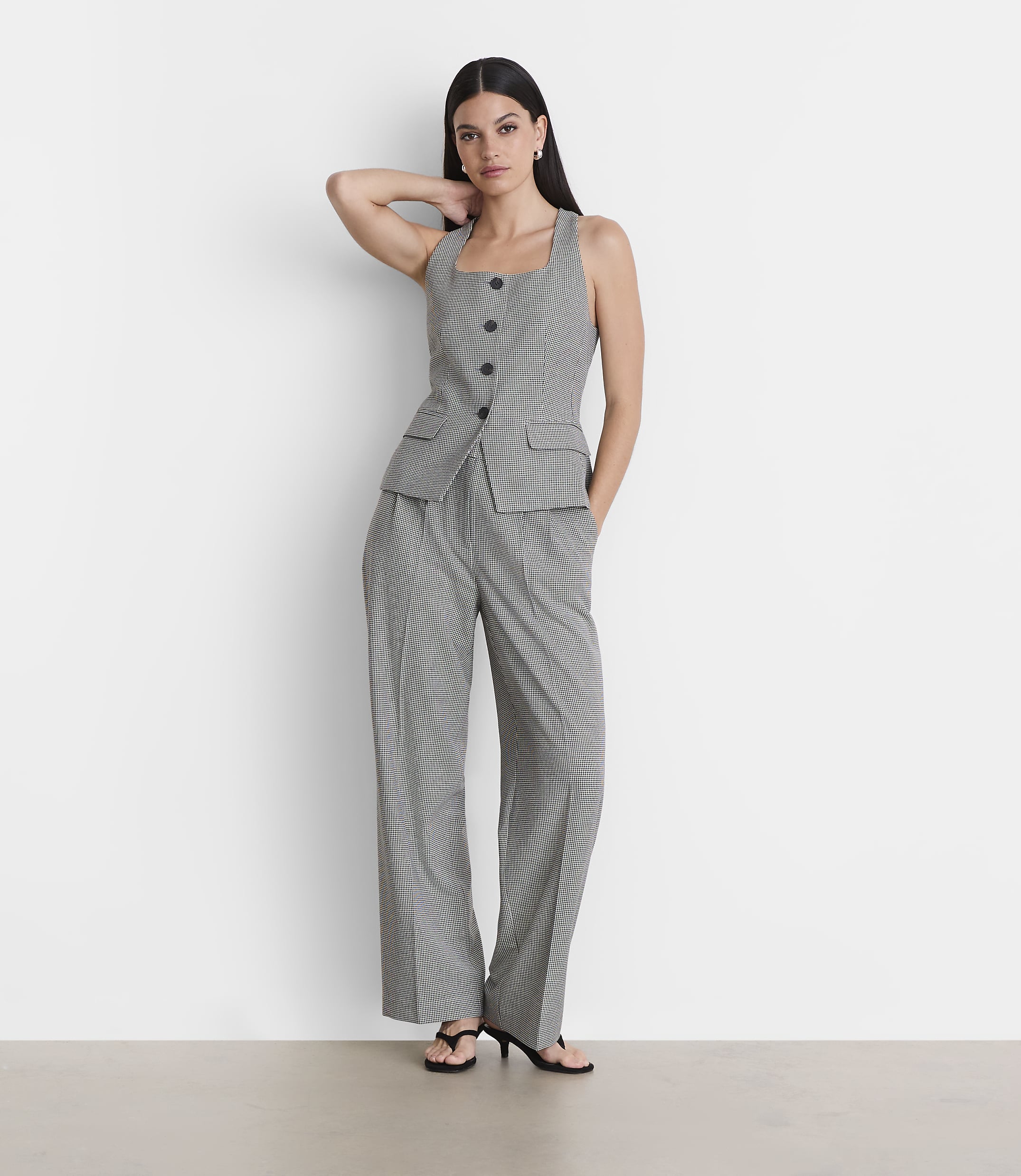 A woman in a checkered vest and pants stands against a plain white wall.