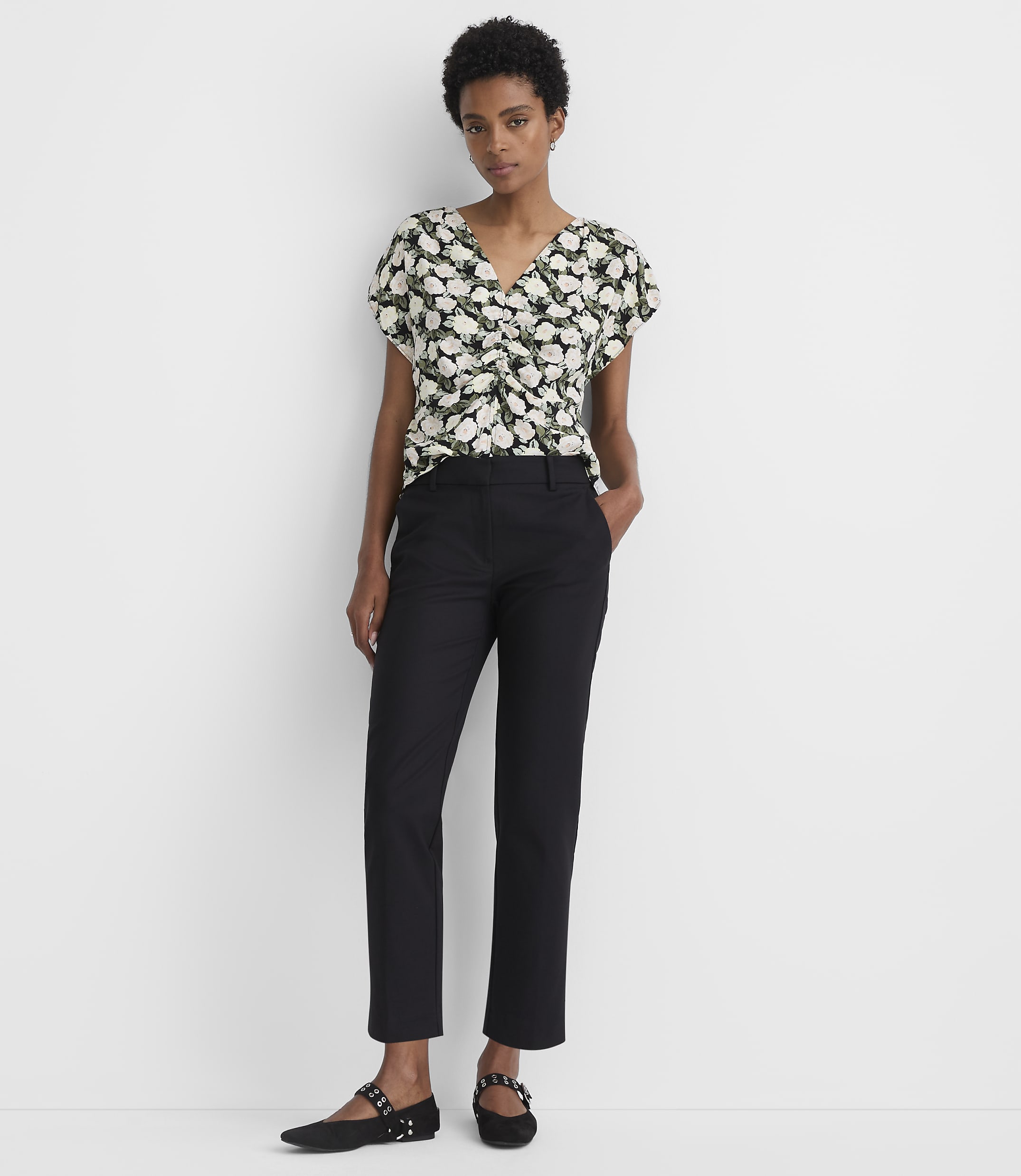 A woman in a floral top and black pants stands against a plain white background.