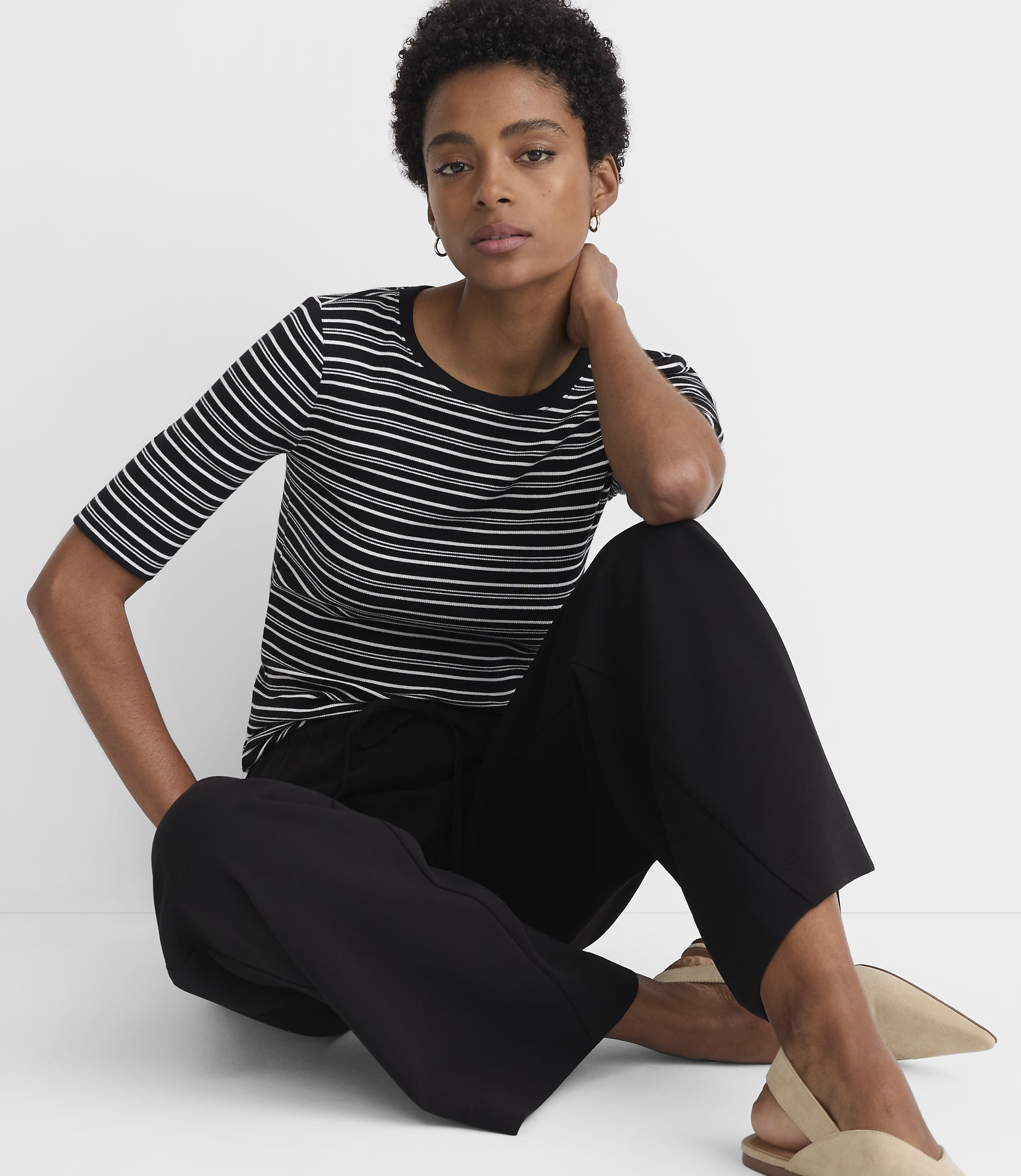 A woman in a striped t-shirt and black pants sits on a plain white background.