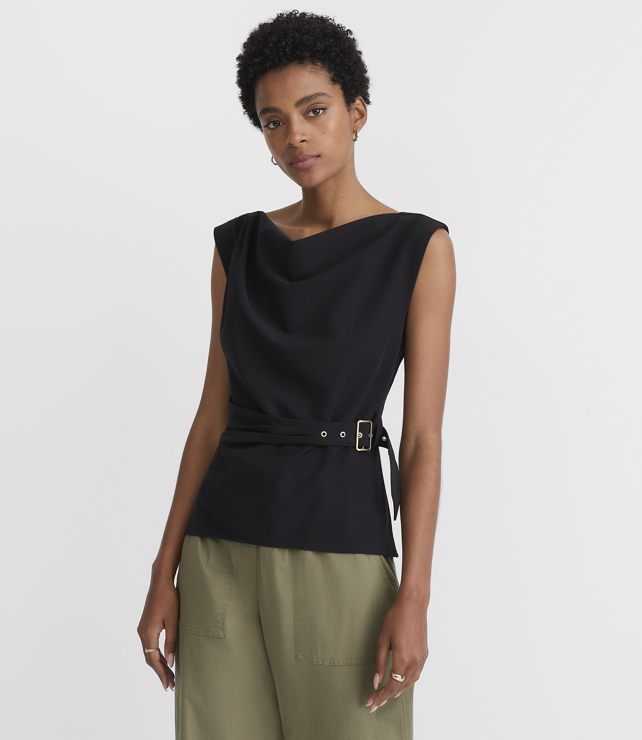 A woman in a black cowl-neck top and olive green pants stands against a plain white background.