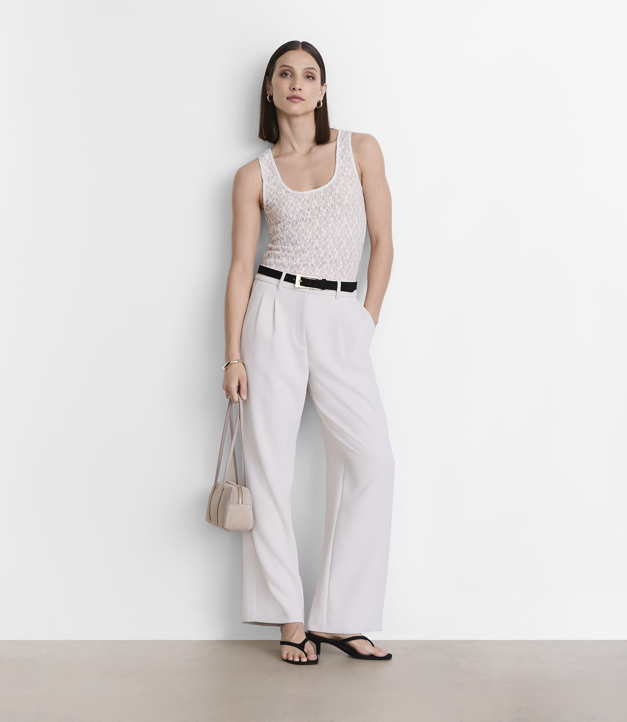 A woman in a white tank top and light-colored pants stands against a plain white wall, holding a small purse.