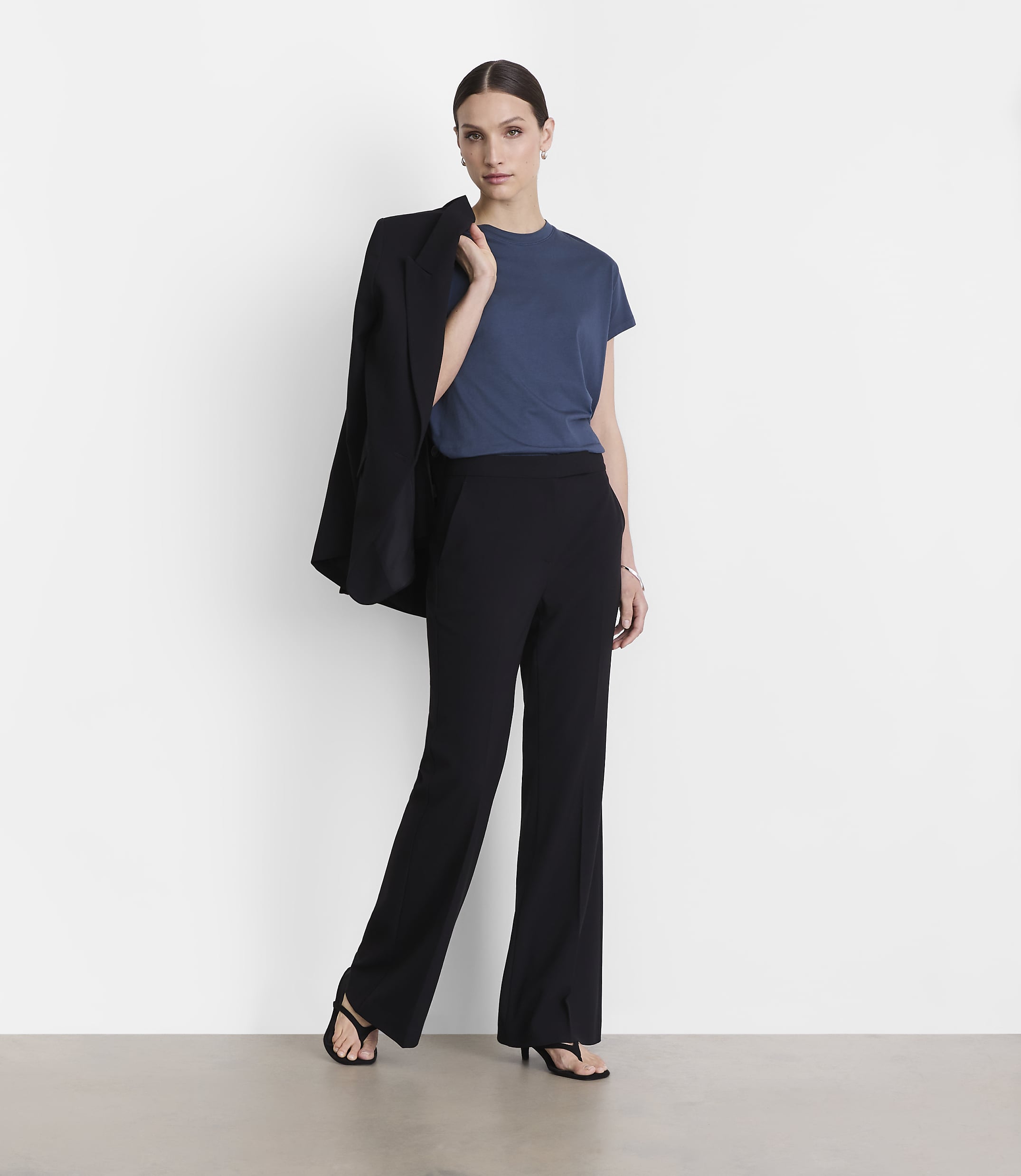 A woman in a blue t-shirt and black pants stands against a white background, with a black blazer draped over her shoulder.