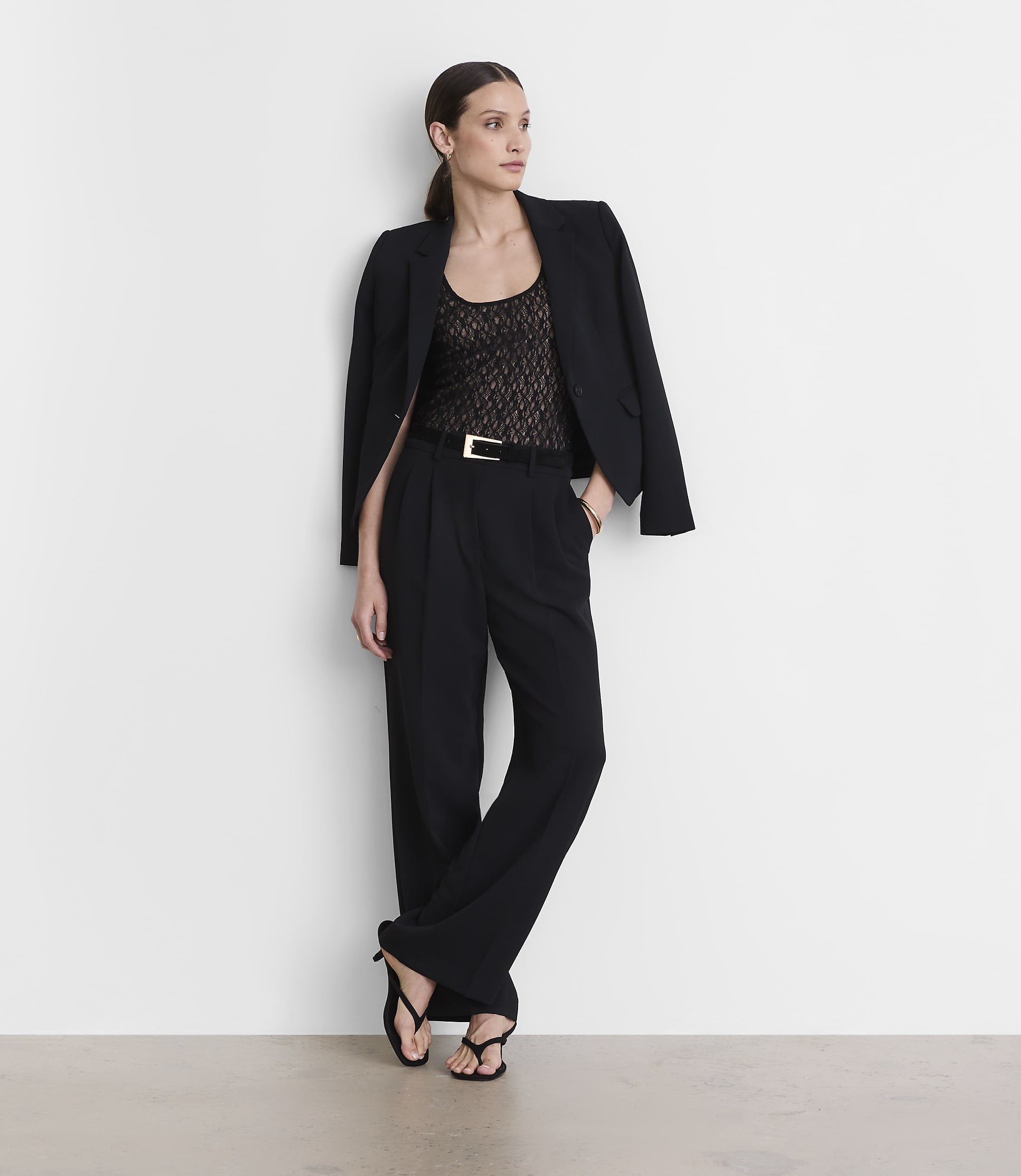 A woman in a black blazer, lace top, and wide-leg pants stands against a plain white wall.