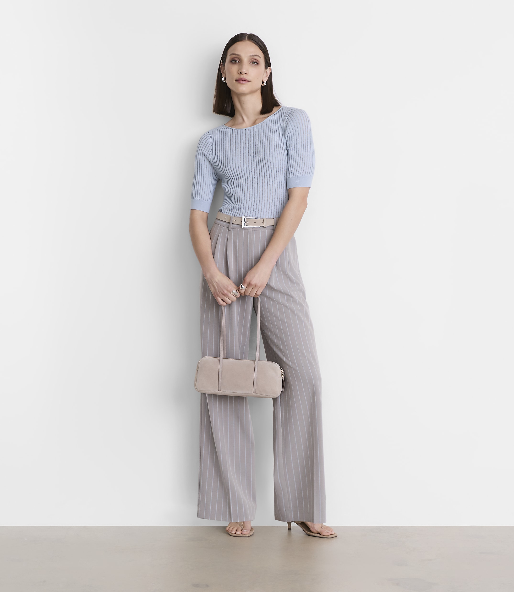 A woman in a light blue ribbed top and pinstripe pants holds a beige clutch in front of a plain white wall.