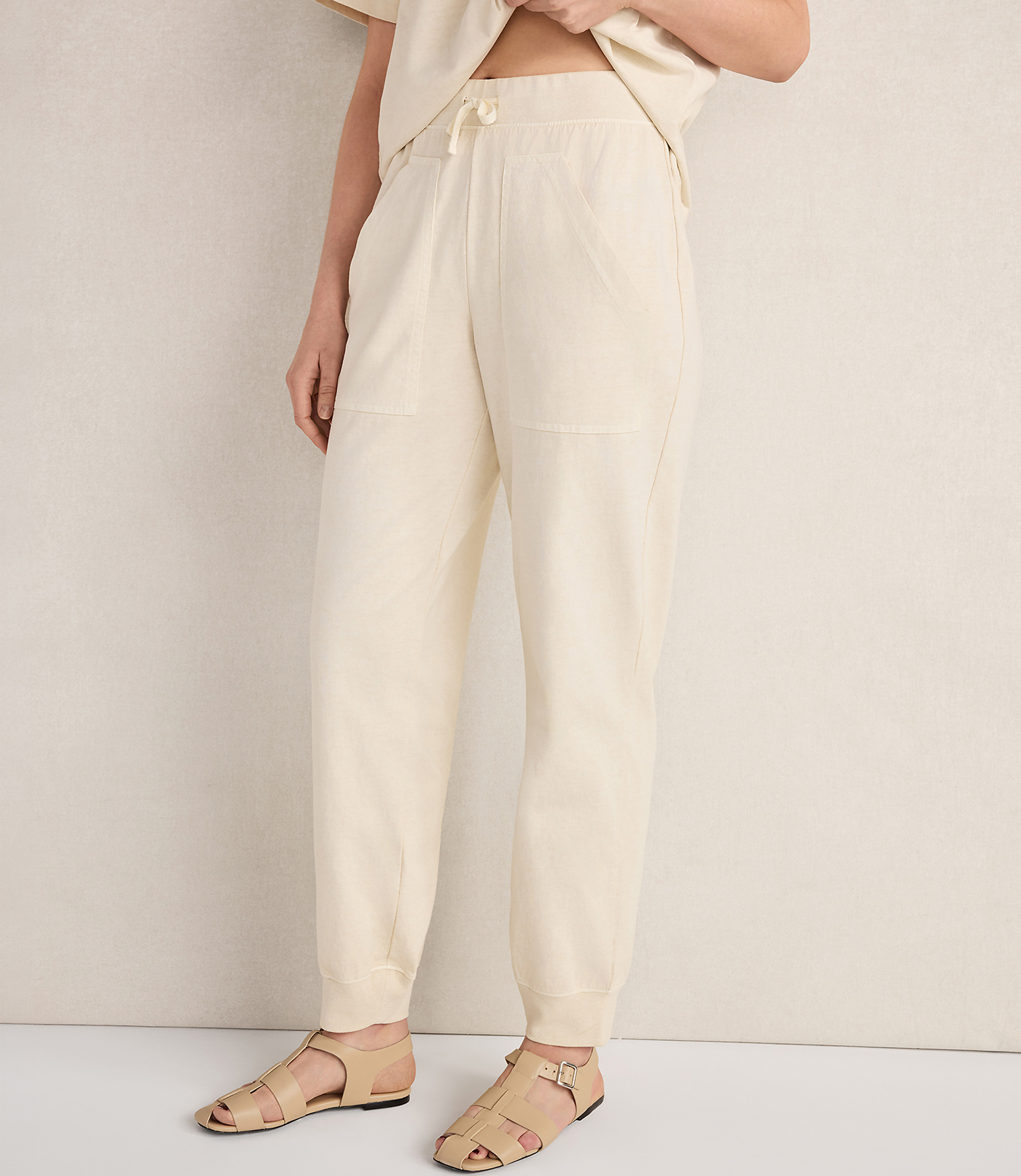A person wearing cream-colored sweatpants and sandals stands against a textured, light-colored wall.