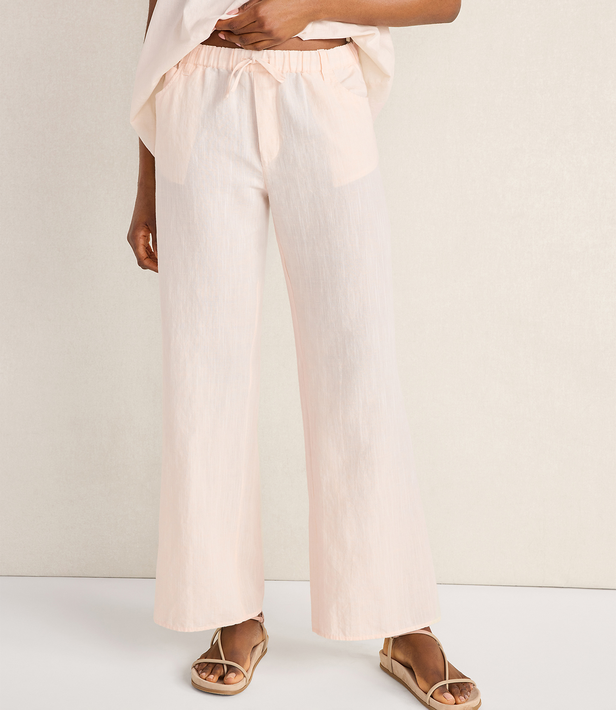 A person wearing light pink wide-leg pajama pants and sandals stands against a plain, light-colored background.